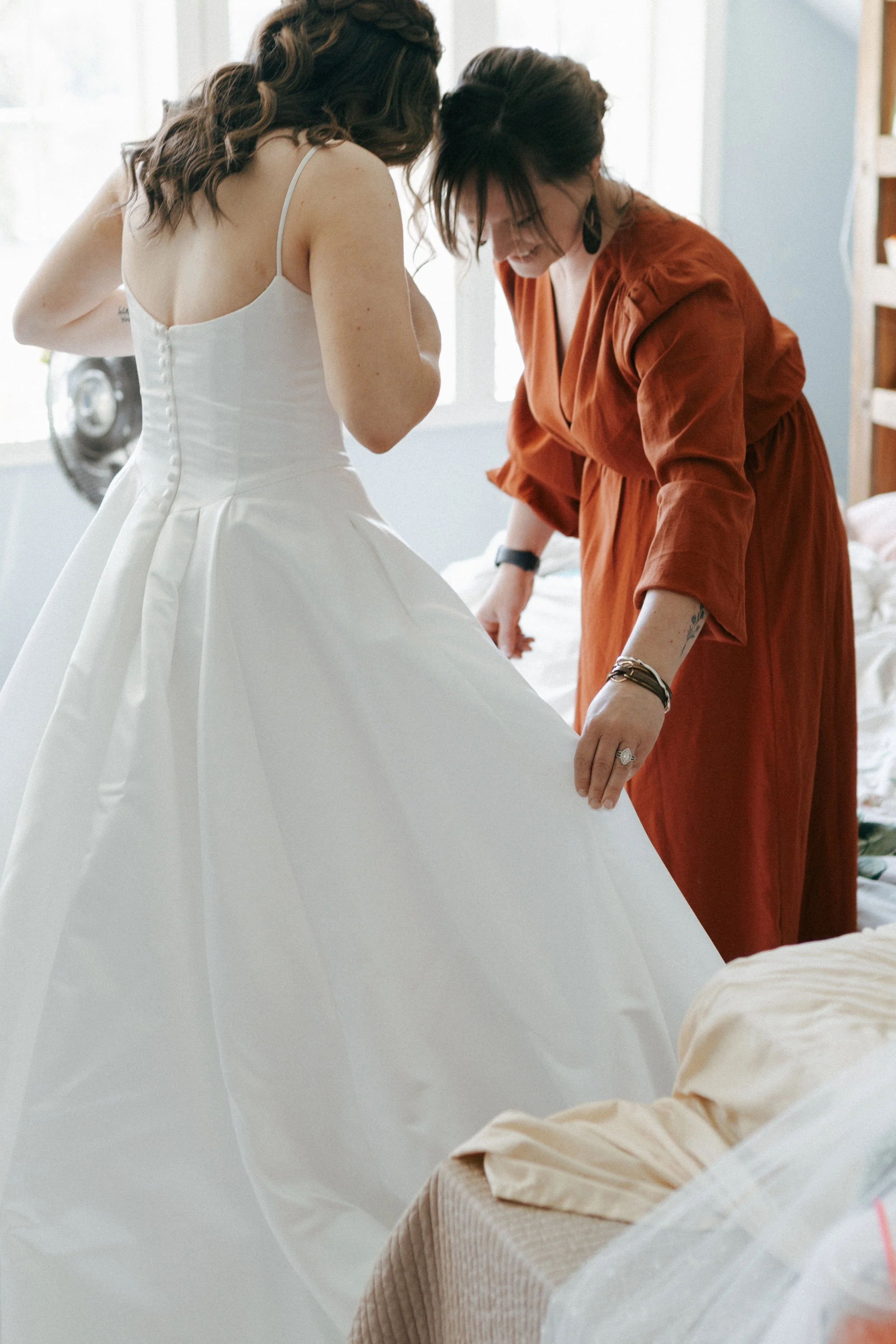 A bride in a white wedding dress being assisted by a woman in an orange dress, as they prepare for a wedding.