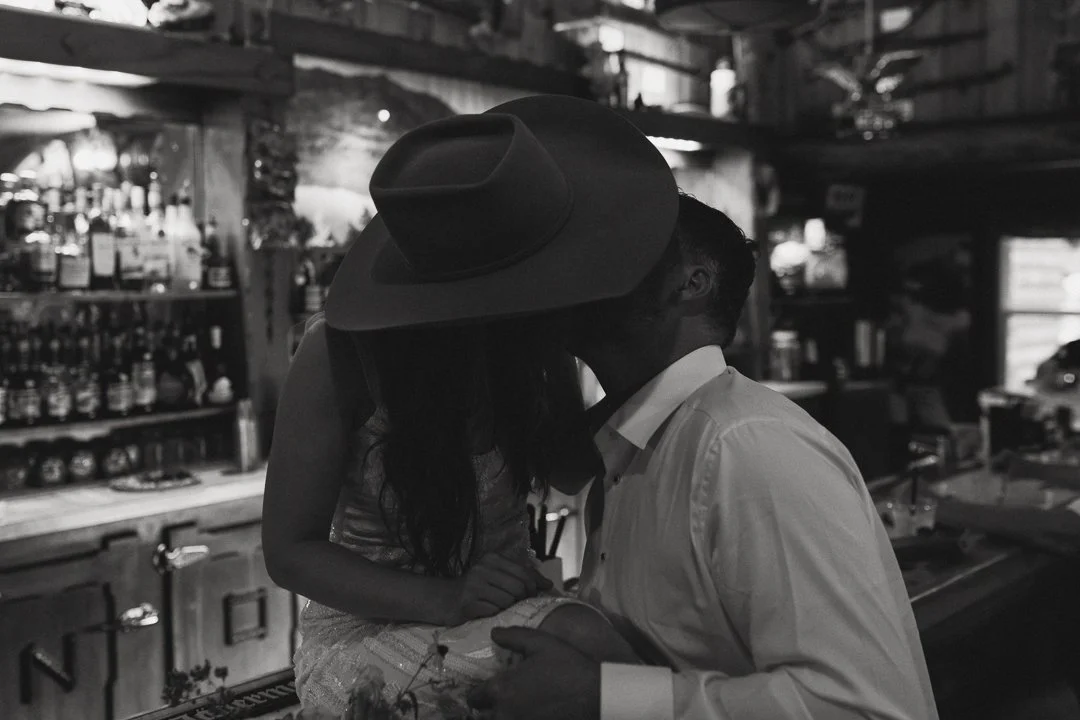 A couple sharing a kiss in a dimly lit bar, with the woman wearing a hat and the man in a white shirt.