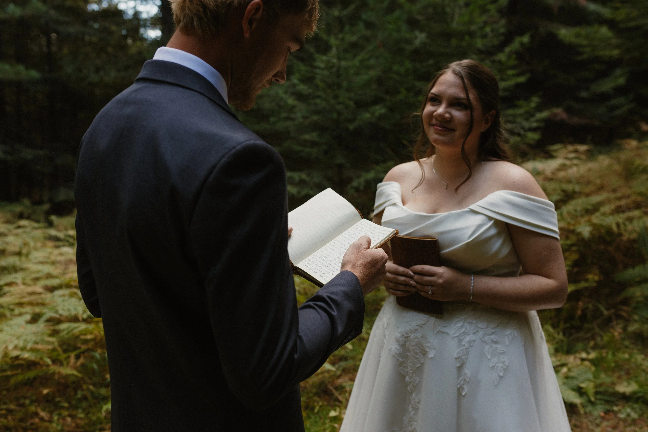 A couple getting married outdoors in a forest, with the groom reading from a notebook and the bride smiling in a white wedding dress, holding a small book.