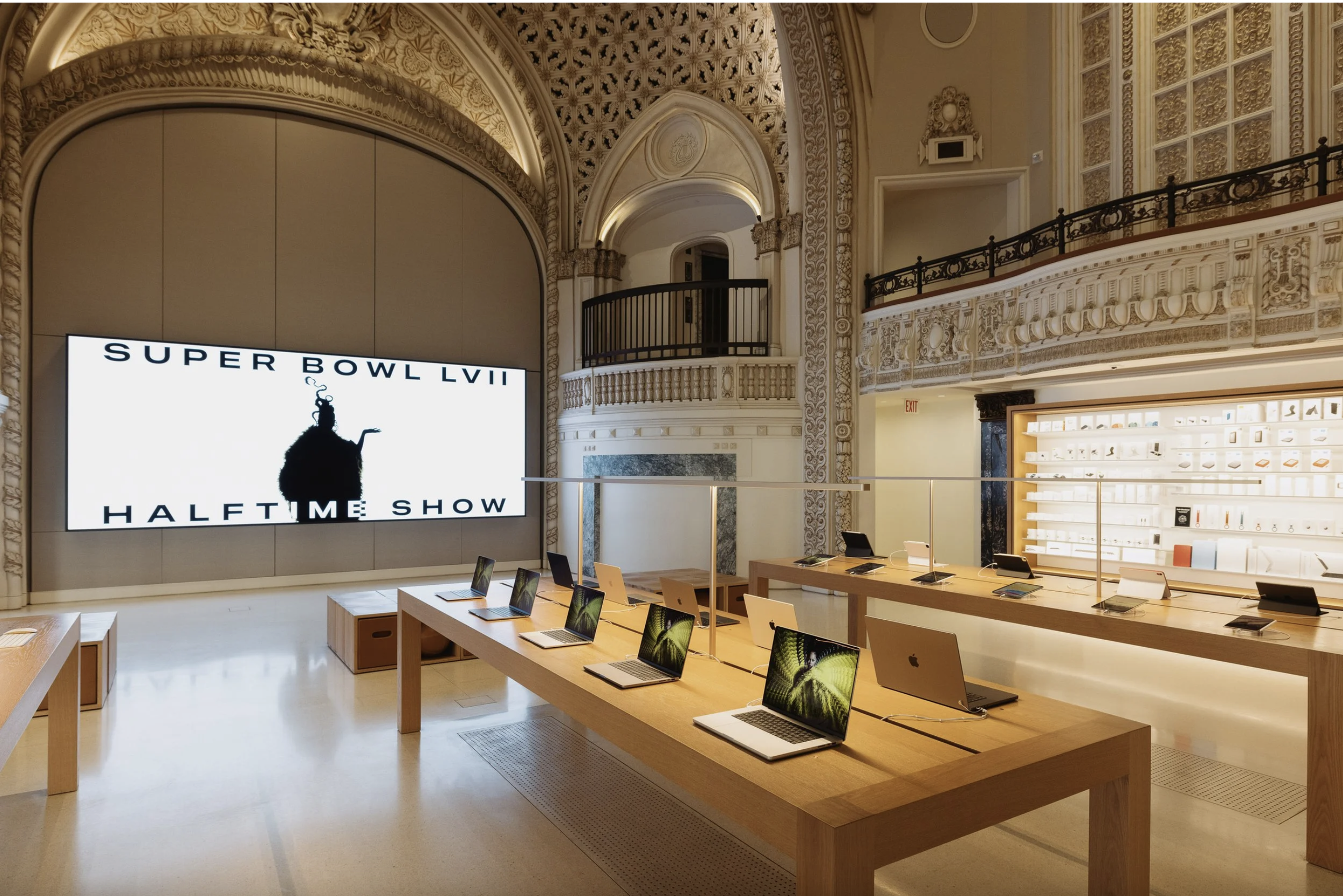 Apple store interior with a large digital screen displaying Super Bowl LII halftime show advertising with Rihanna, wooden tables with laptops, and a decorative, ornate interior design.