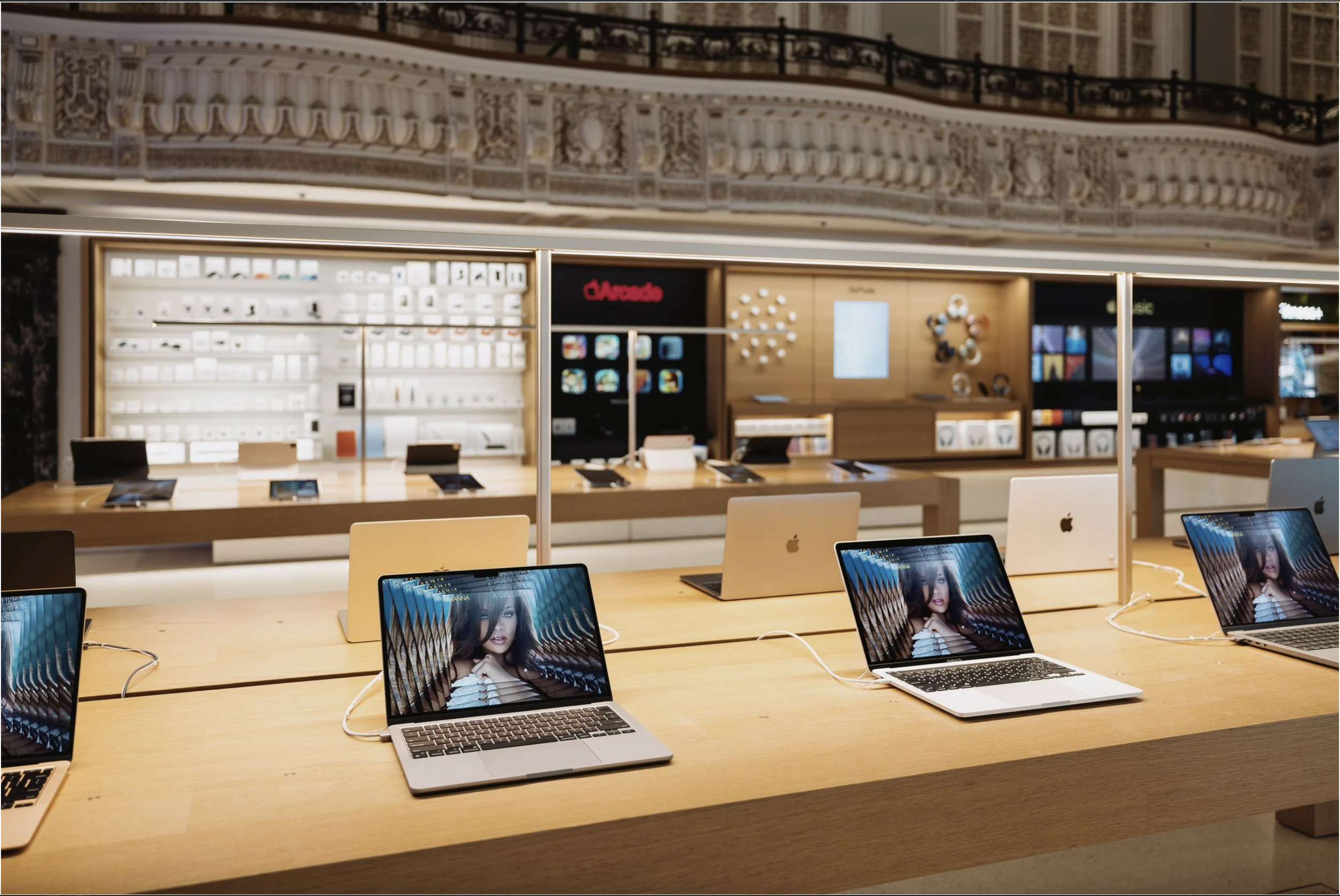 Inside an Apple Store, displays of MacBook laptops on a wooden table with screens showing a Rihanna's portrait, in front of a background with shelves of accessories and gadgets. Apple Music Spatial Audio