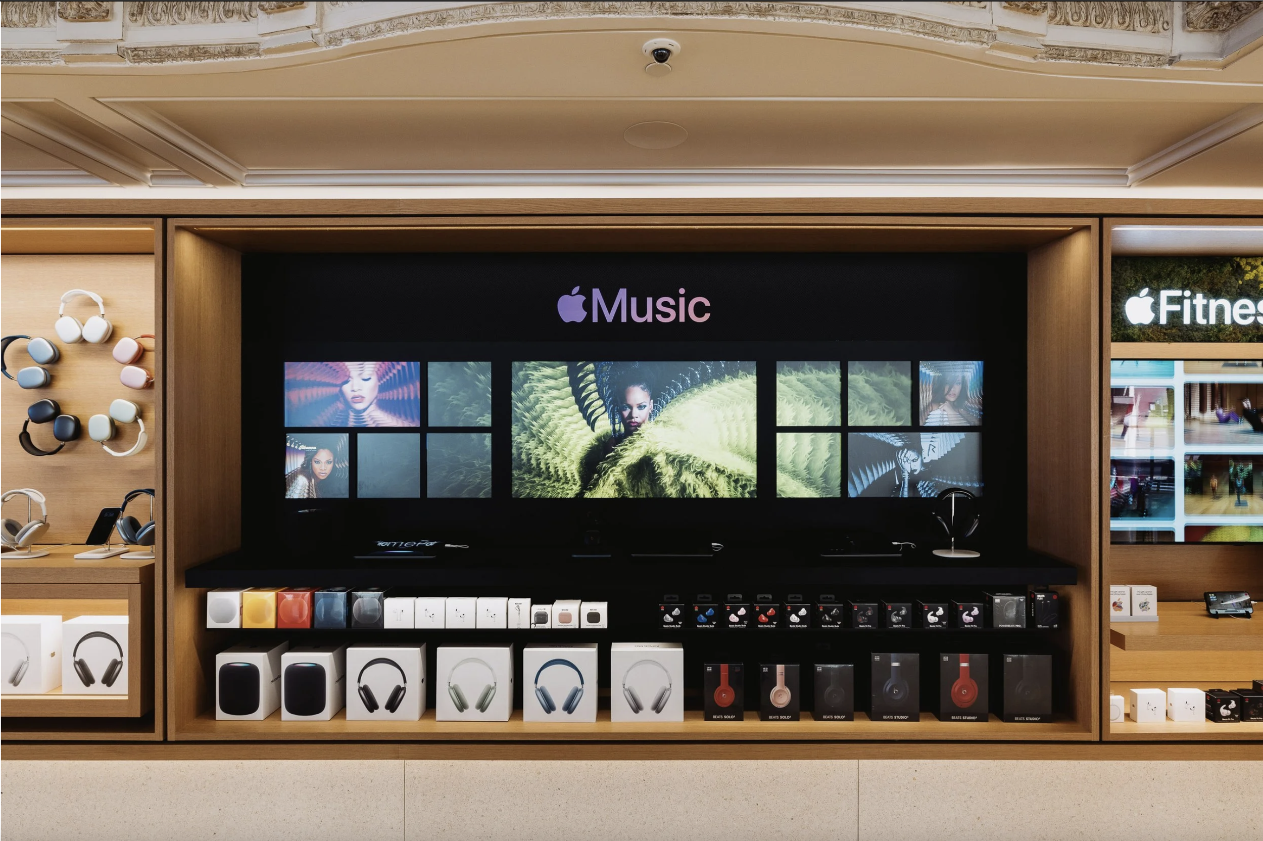Display of Apple-branded headphones, speakers, and accessories in a retail store with a digital screen showing music-related images. Rihanna Spatial Audio Apple Music