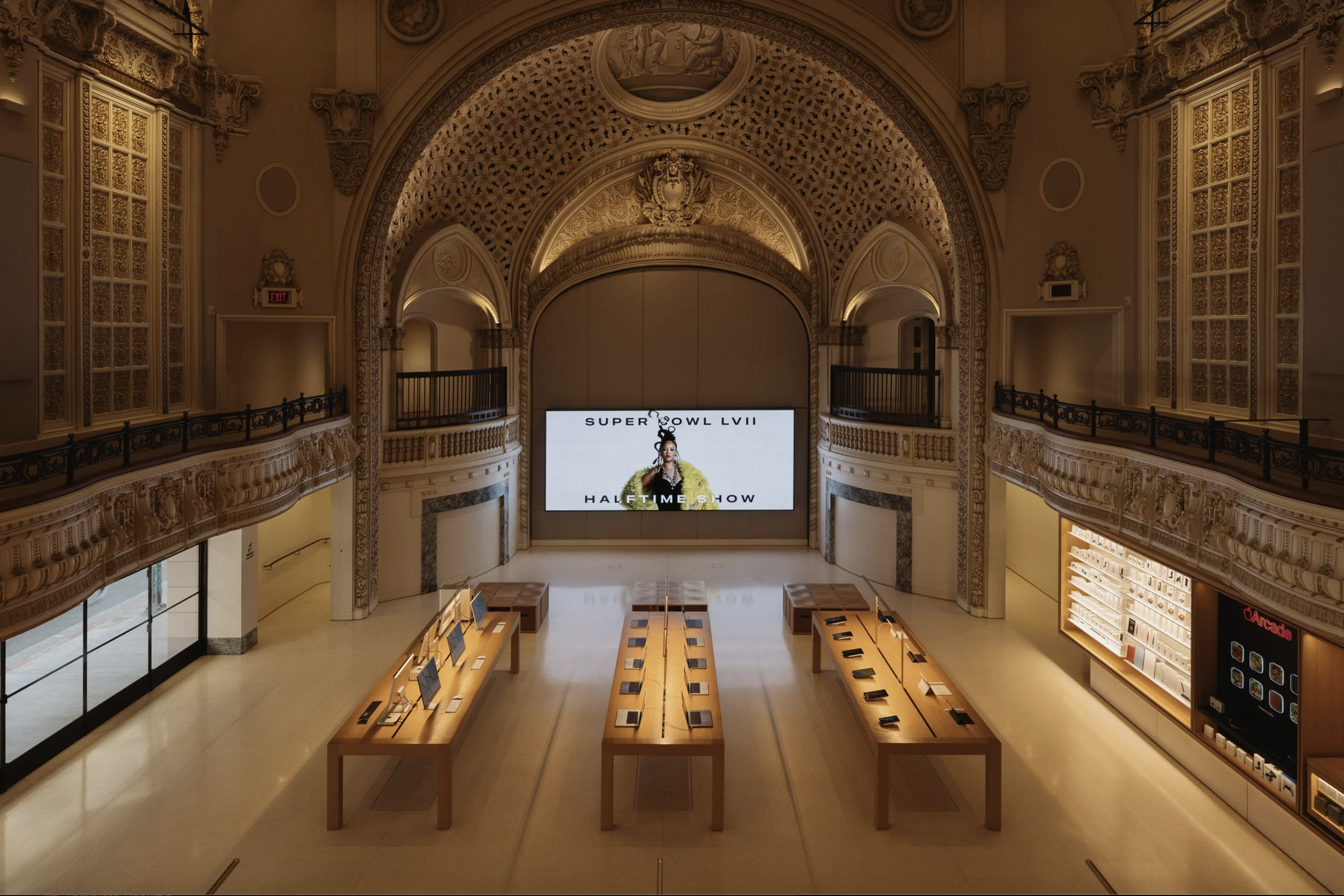 Empty Apple Store with wooden tables displaying iPads, a large digital screen with a Rihanna and text referencing the Super Bowl LVII halftime show, ornate gold and white architectural details, and a high ceiling.