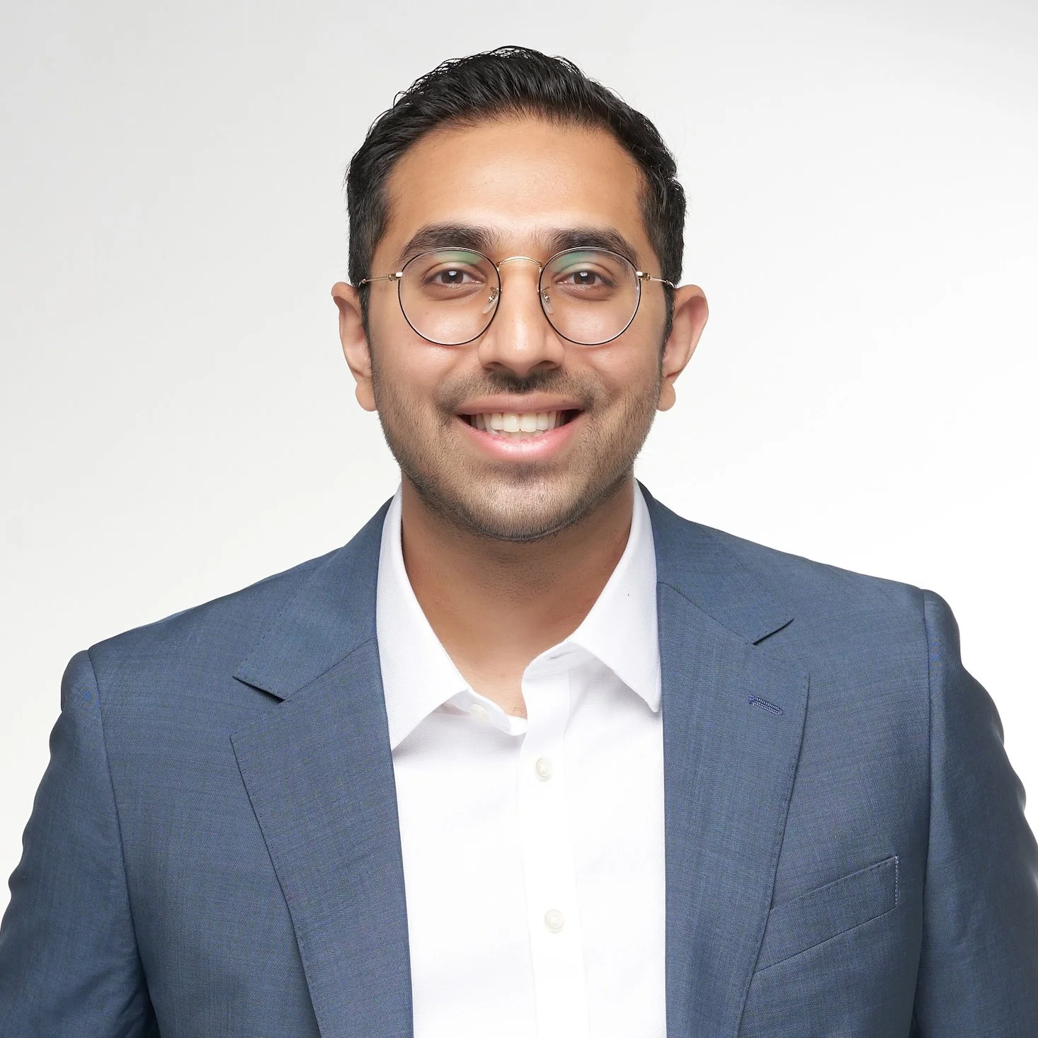 Professional headshot of a young man in a blue suit with glasses, smiling against a plain white background.