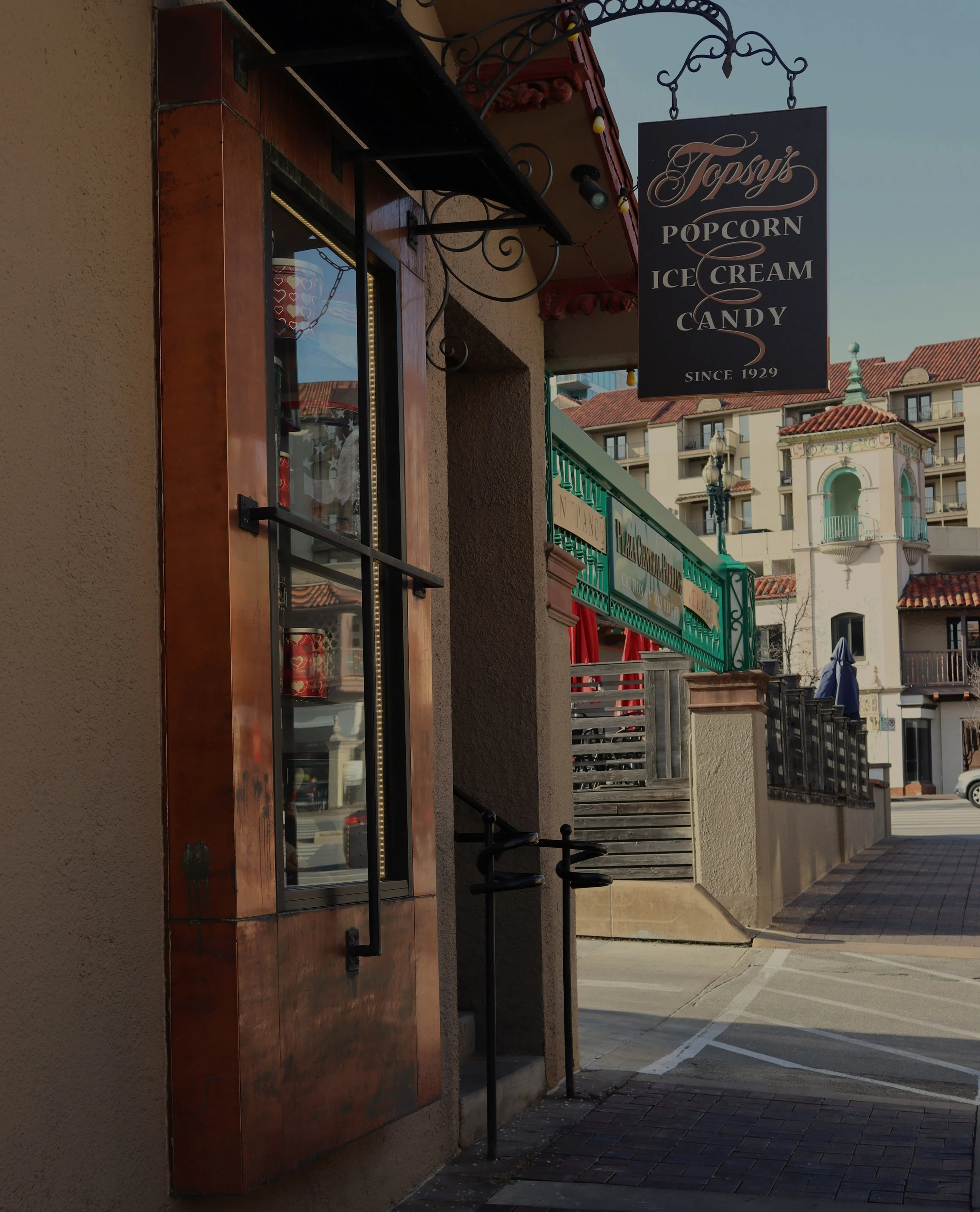 Sign for Topsy's popcorn, ice cream, and candy shop outside a building with a window and outdoor seating, in a town with multi-story buildings featuring Spanish-style tile roofs.