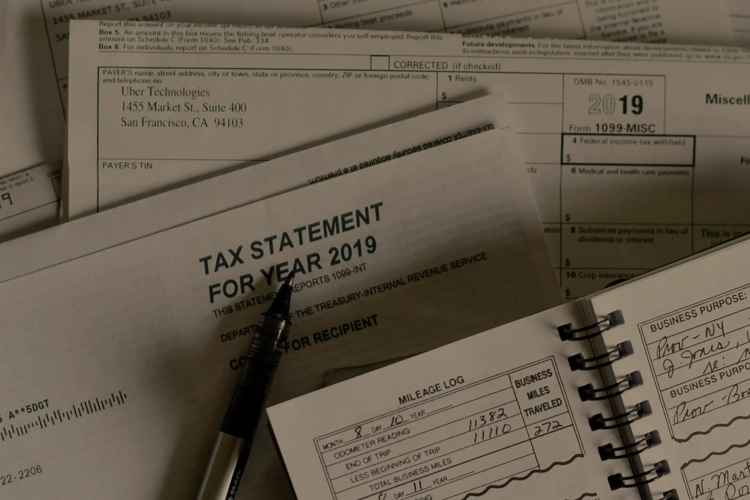 Close-up of various documents including a 2019 tax statement, a mileage log, and a business planner with handwritten notes, a pen resting on the tax statement.