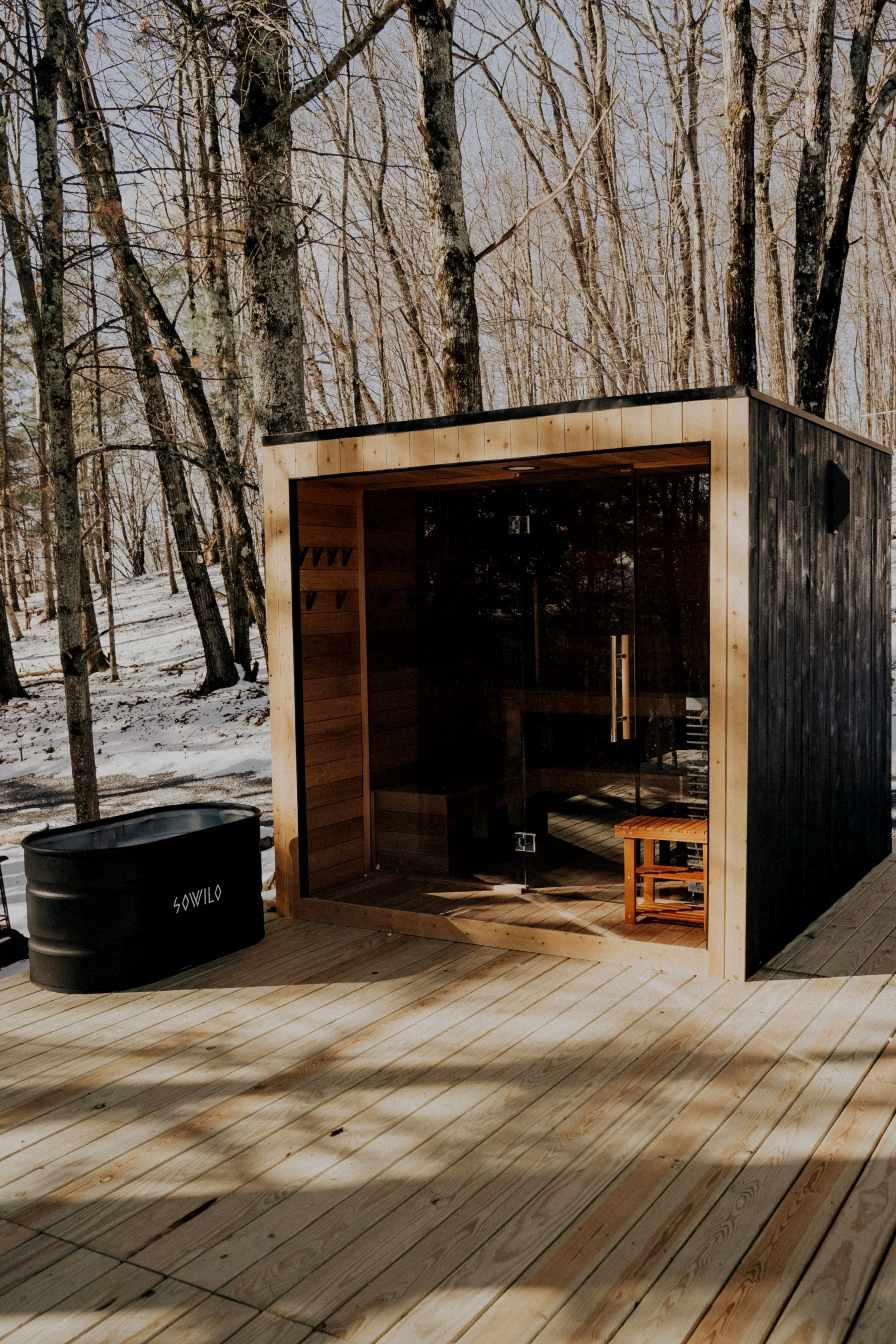 A small wooden sauna with glass door situated on a wooden deck in a snowy forest.