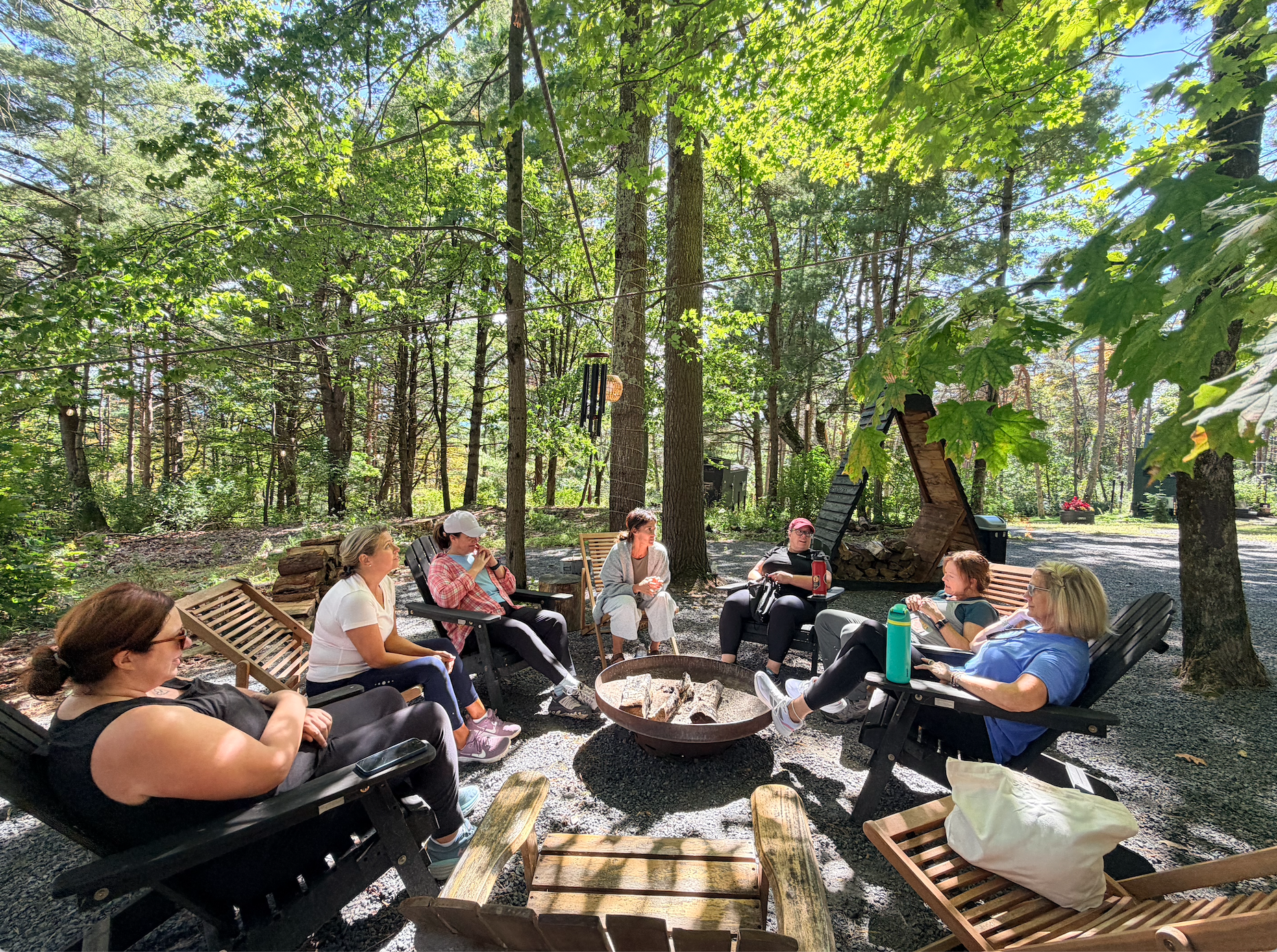Group of women sitting in Adirondack chairs around a fire pit in a wooded outdoor setting, engaging in conversation during daytime.