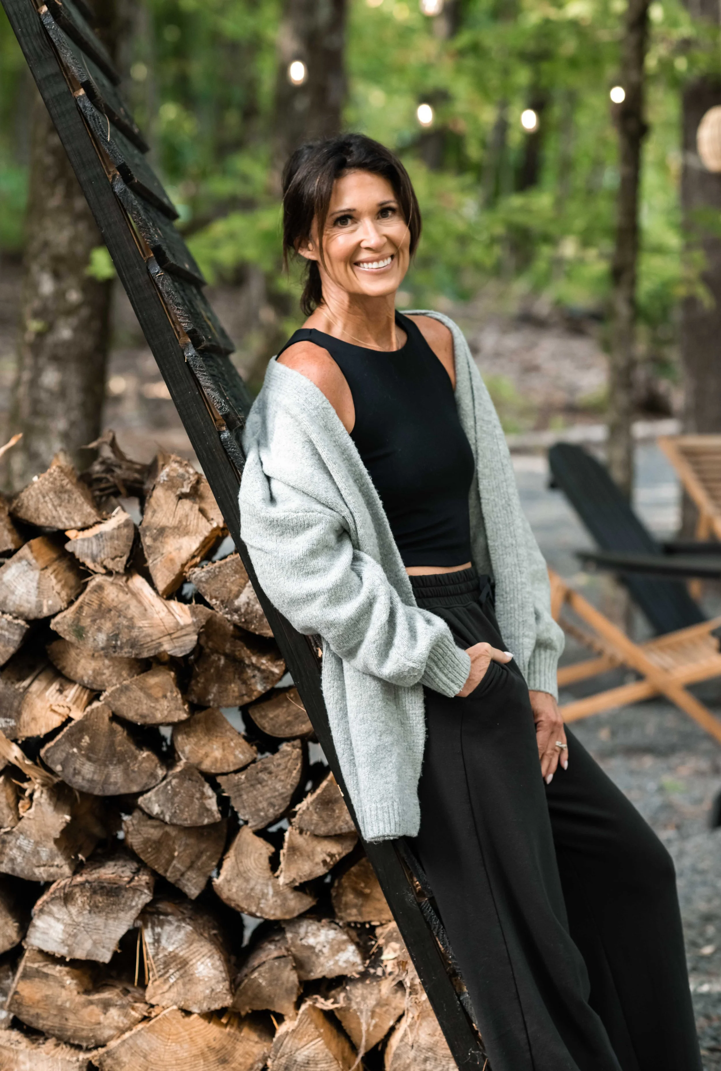 A woman smiling, wearing a black tank top and a gray cardigan, leaning against a woodpile in a forested area with chairs and string lights in the background.