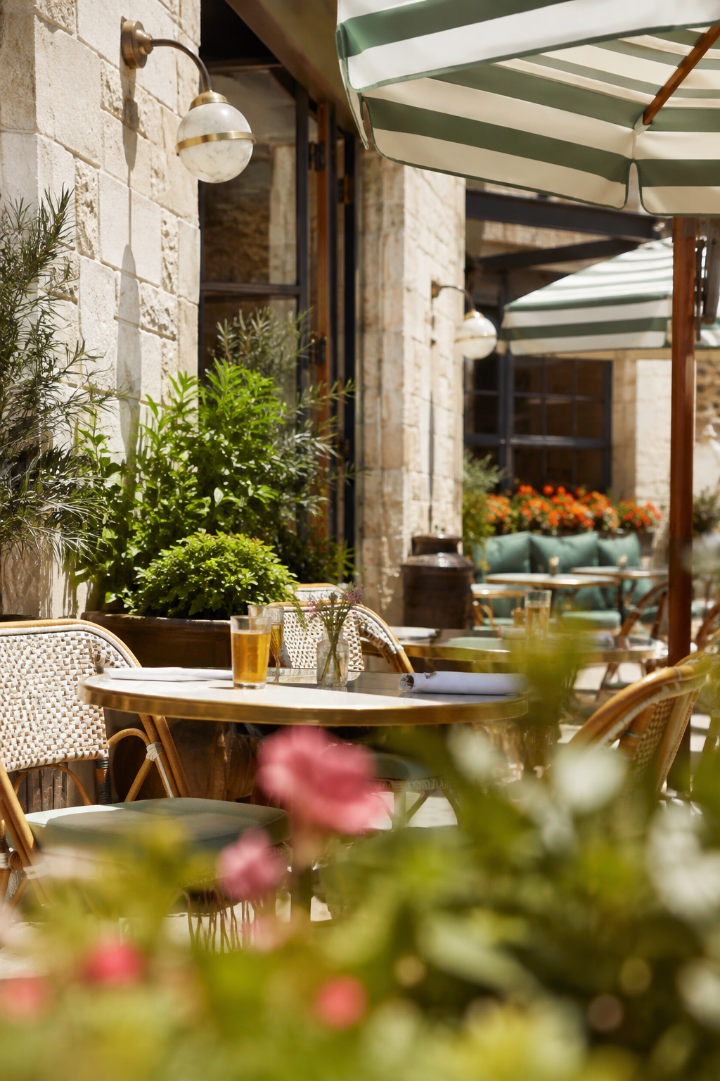 An outdoor patio of a restaurant or cafe with round tables, wicker chairs, and green umbrellas. There are potted plants and flowers, including pink blossoms in the foreground, and a stone wall with windows in the background.