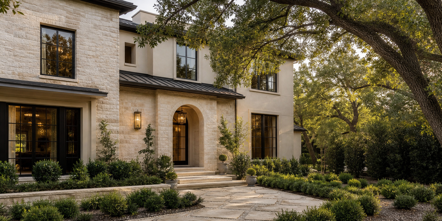 Front view of a modern house with stone and stucco exterior, large windows, a black metal roof, and a landscaped garden with trees, bushes, potted plants, and a stone pathway at sunset.