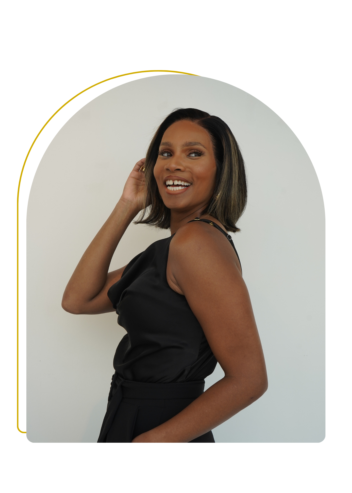 A woman with shoulder-length black hair wearing a black sleeveless top and high-waisted pants, smiling and touching her hair, standing against a plain white background.