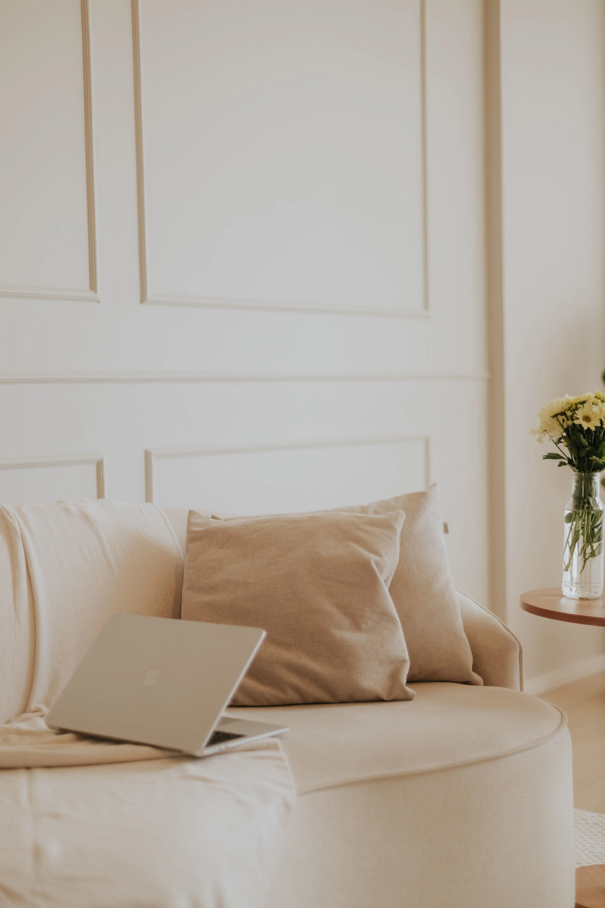 A beige sofa with matching pillows in a bright, minimalist living room. A silver laptop is open on the sofa and a small round wooden side table holds a glass vase with white flowers.