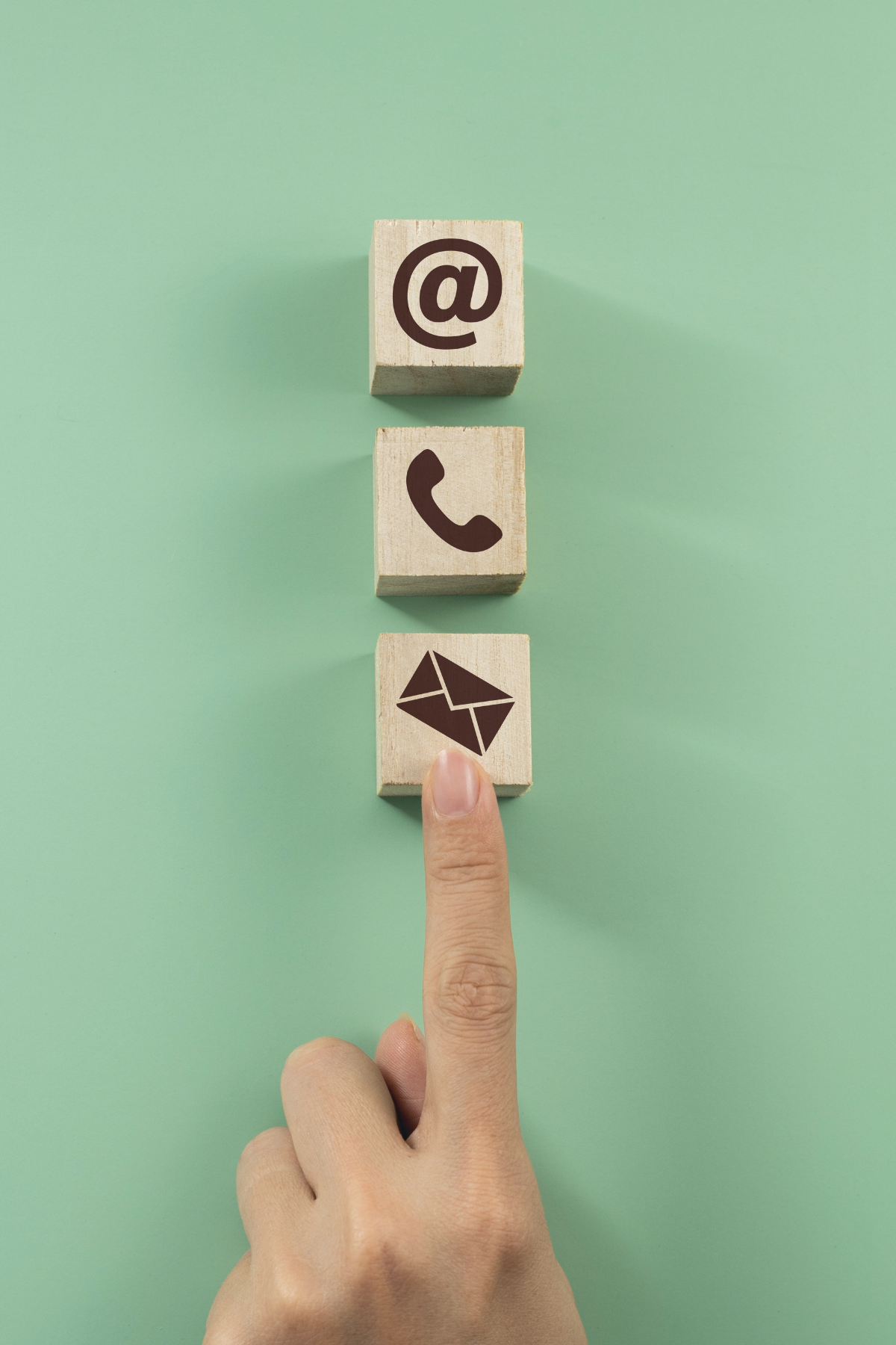 Three wooden blocks with communication icons: email at the bottom, a phone in the middle, and a social media at the top, with a finger pointing at the email icon.