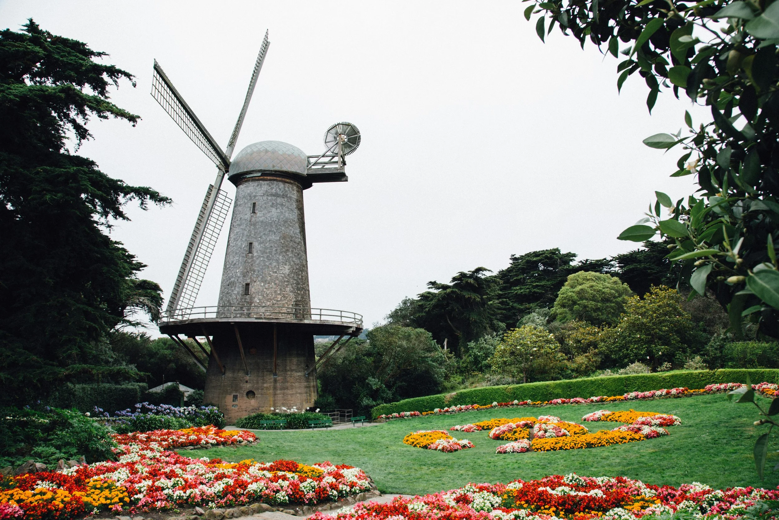 A tall windmill structure with four large blades surrounded by a landscaped garden with colorful flowers and green trees.