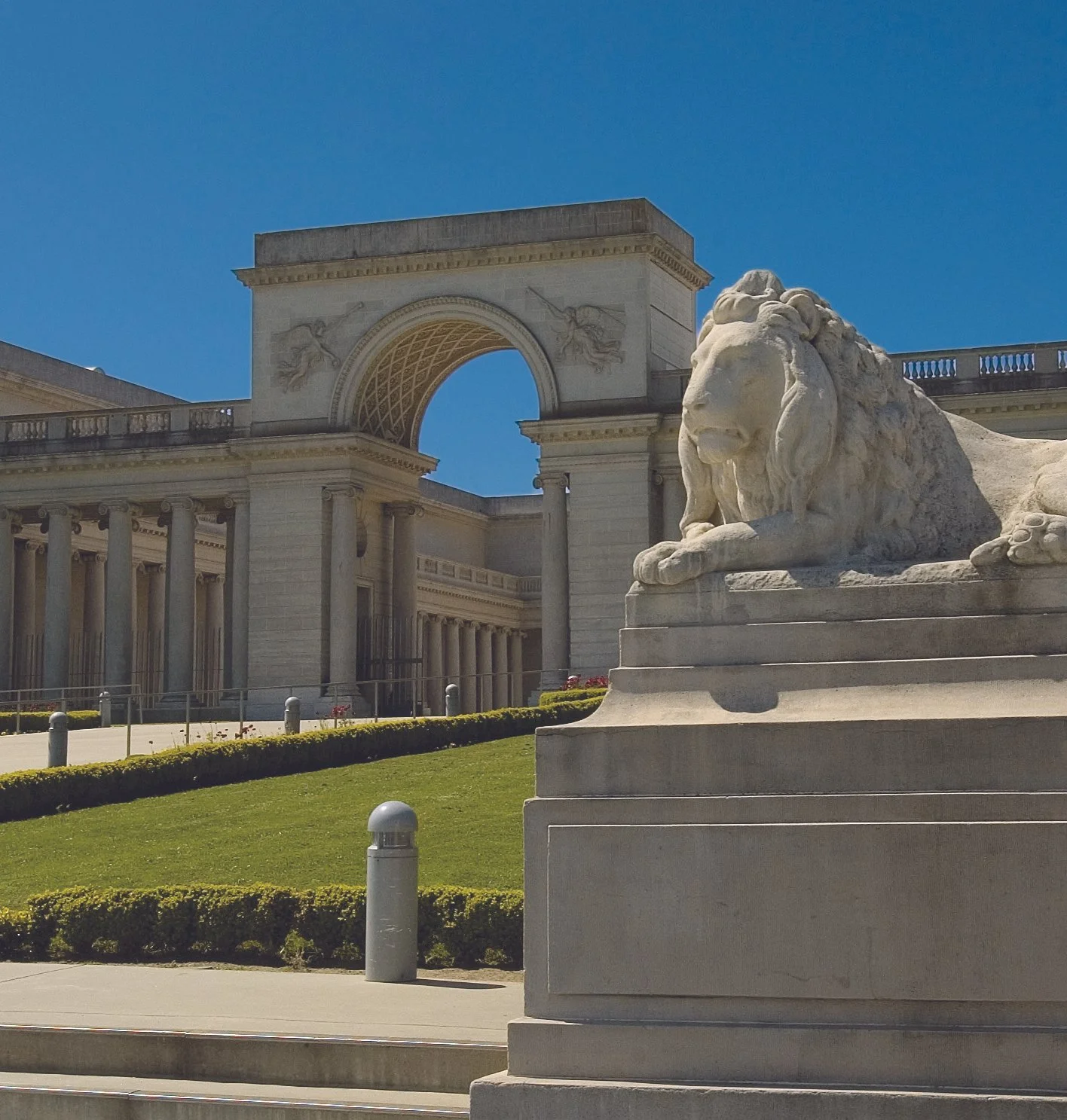 Stone statue of a lion lying down in front of a neoclassical building with an arch and columns under a clear blue sky.