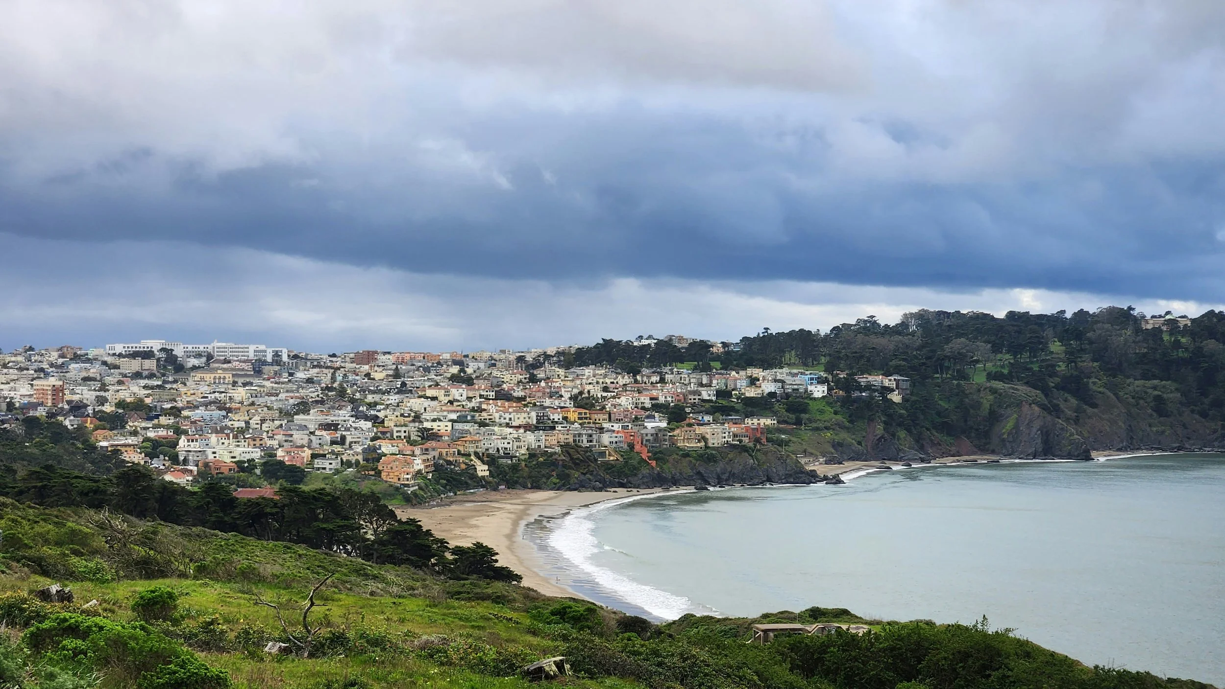 Coastal cityscape with hills, residential buildings, and beach under a cloudy sky.