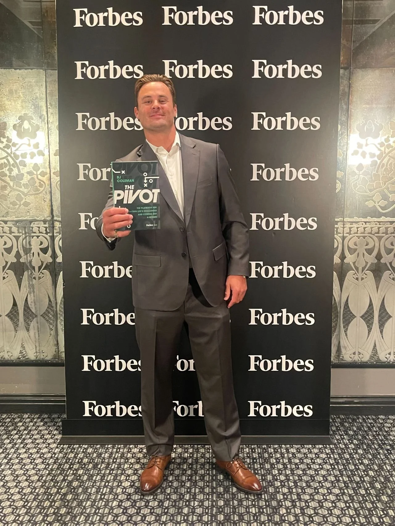 A man in a gray suit holding a book titled 'The Pivot' in front of a black backdrop with multiple 'Forbes' logos.
