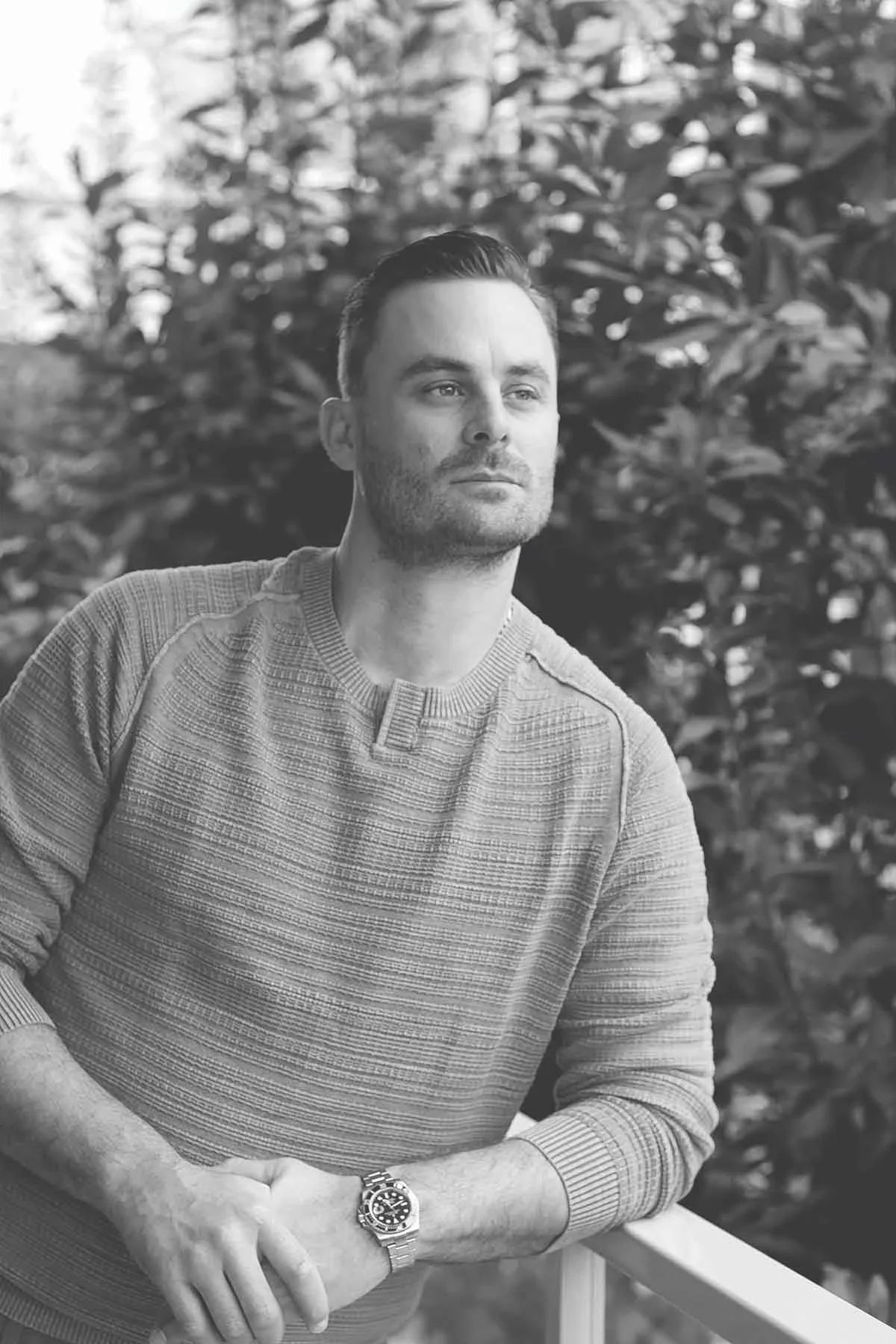 A man with dark hair and a beard, wearing a light sweater, looks thoughtfully to the side while standing outdoors near a railing, with trees in the background.