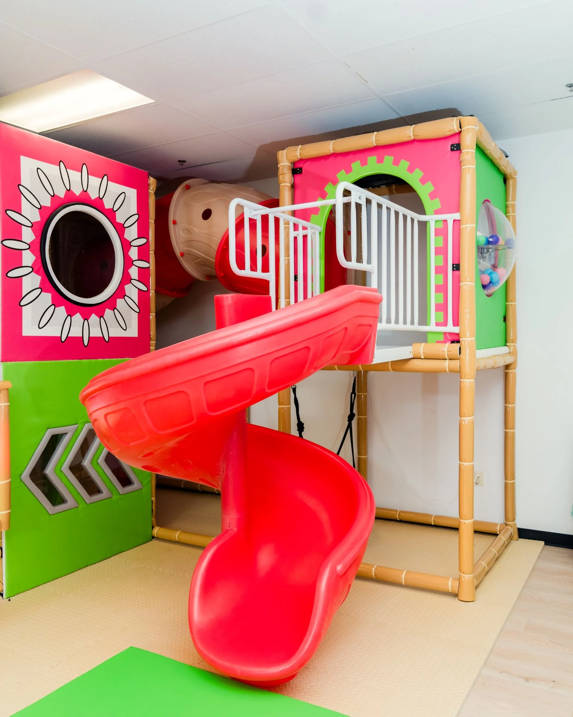 Indoor playground with a red spiral slide, pink climbing structure with netted areas, and colorful play panels with geometric designs.