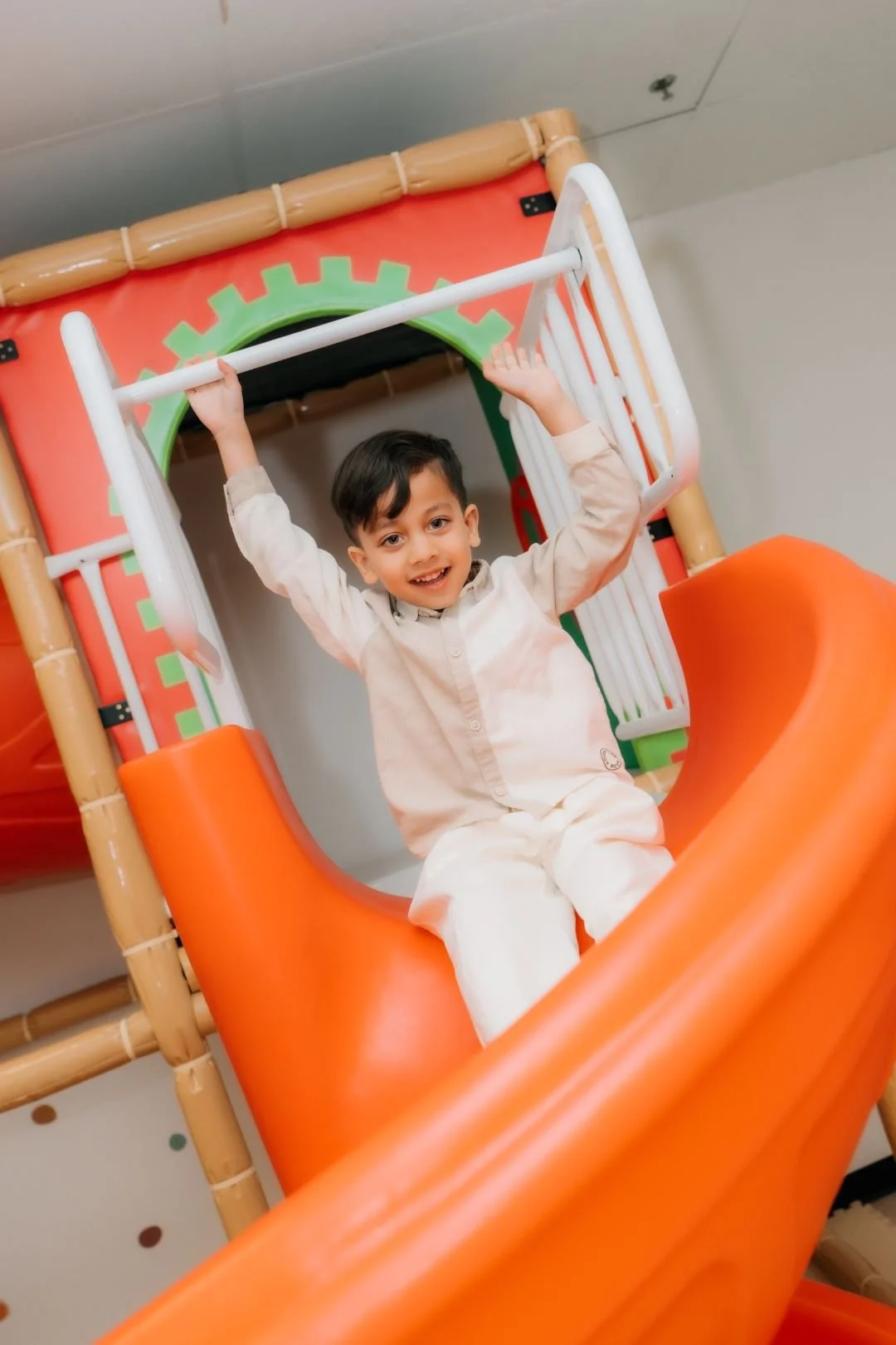 A young boy with dark hair smiling as he sits at the top of an orange play slide, holding onto the white railings, in an indoor play area.