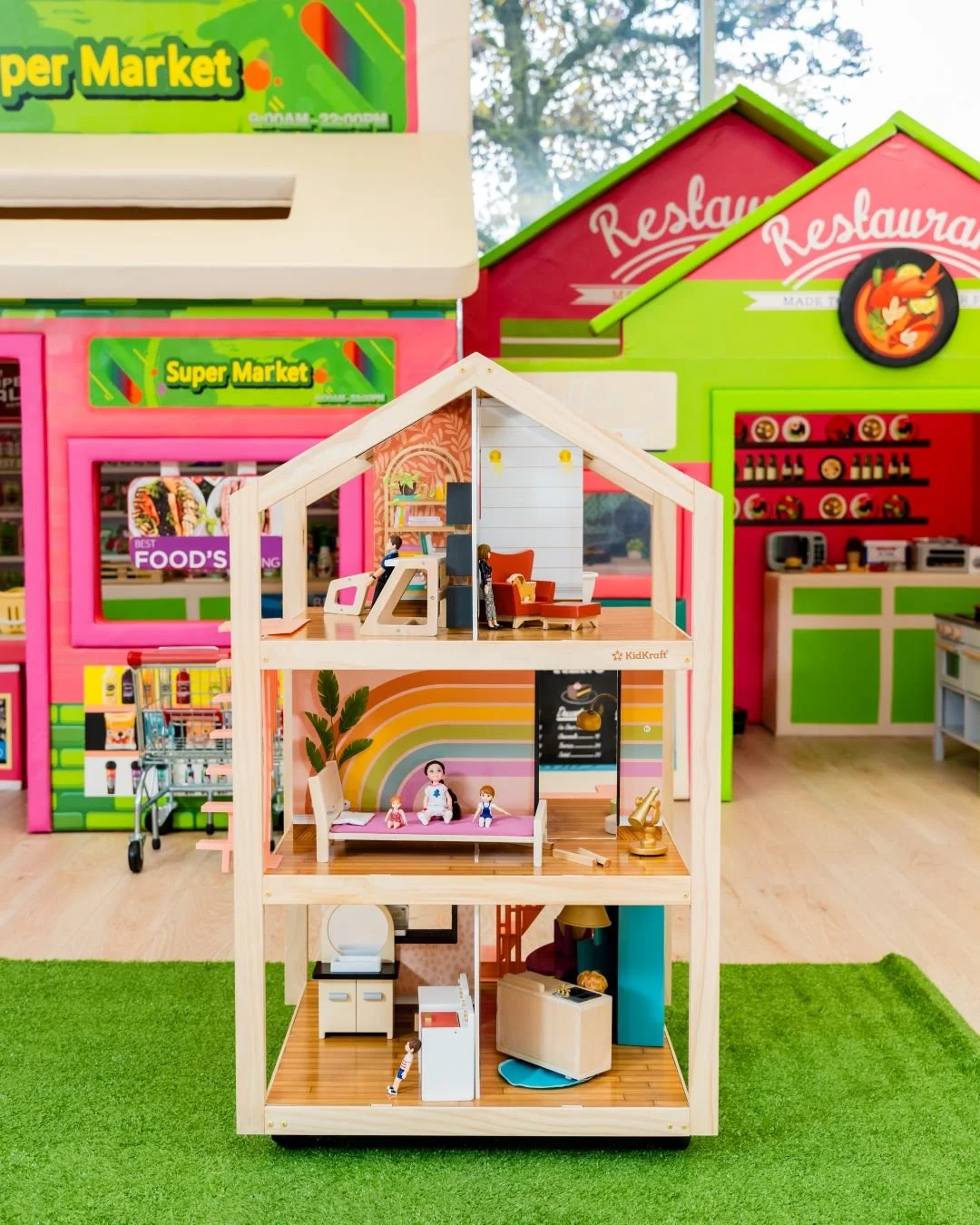 Colorful multi-story toy dollhouse with miniature furniture and dolls inside, set in a play area with a green carpet and a retail store background.