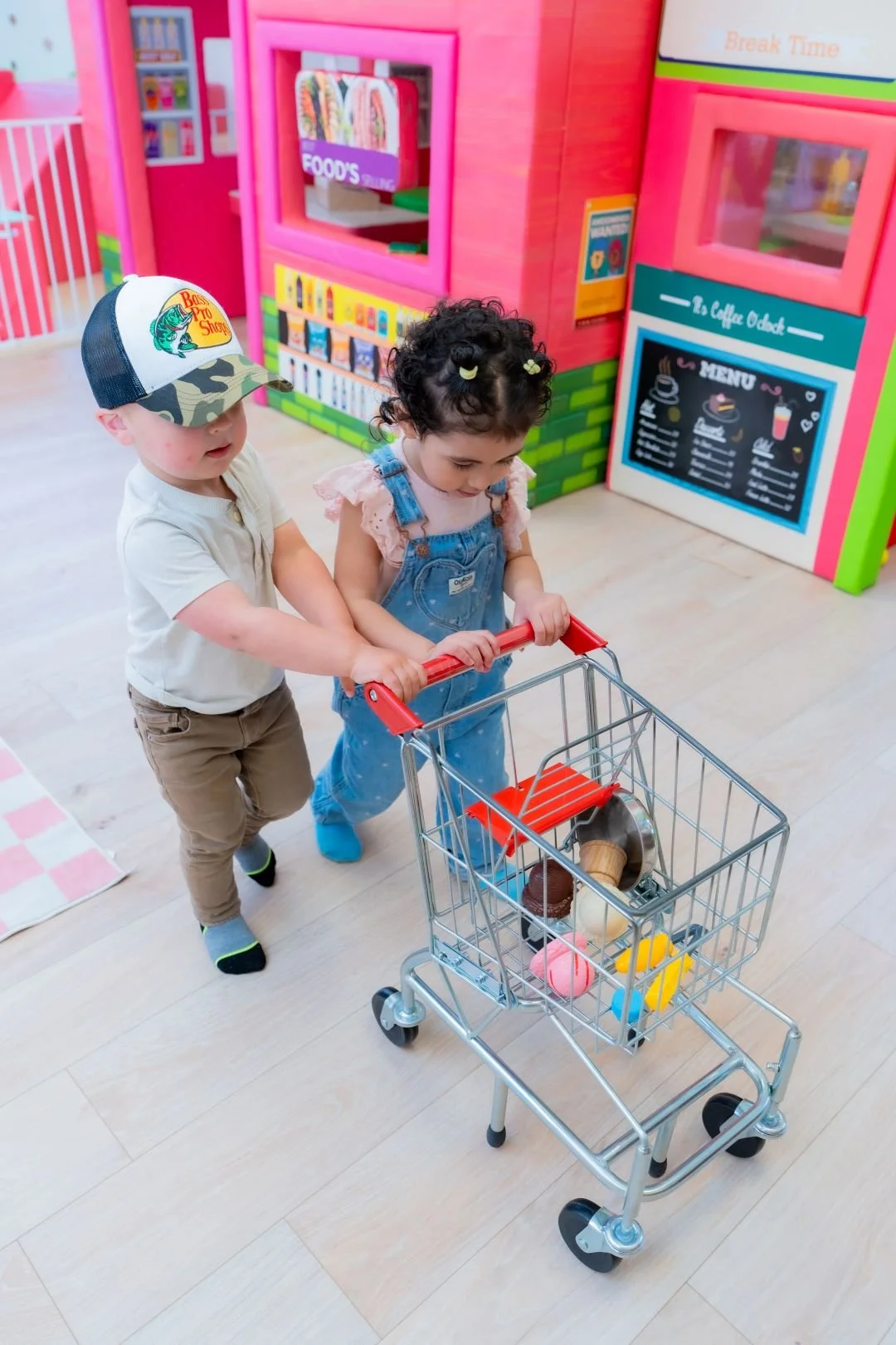 Two young children, a boy and a girl, pushing a toy shopping cart inside a colorful play area resembling a supermarket, with pink and green decor and a menu board in the background.