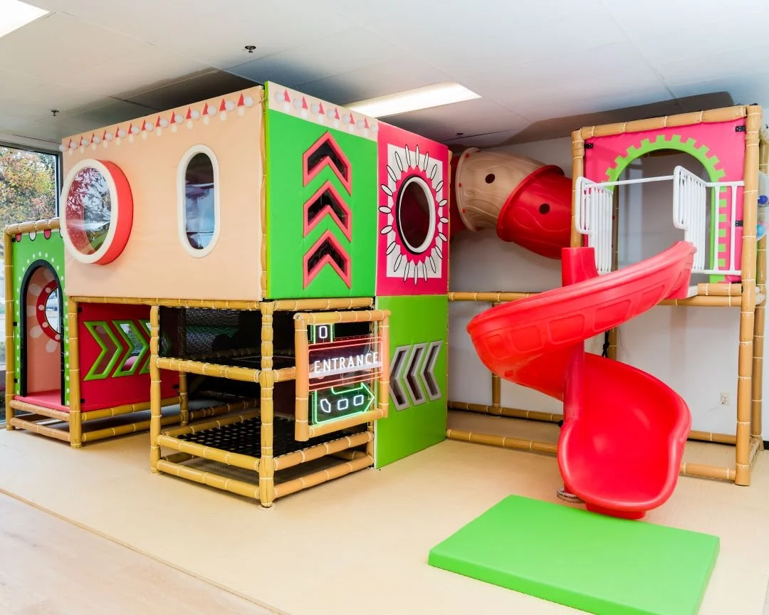Indoor children's playground with colorful play structures, a red spiral slide, and a tunnel, surrounded by bright green, pink, and beige panels.