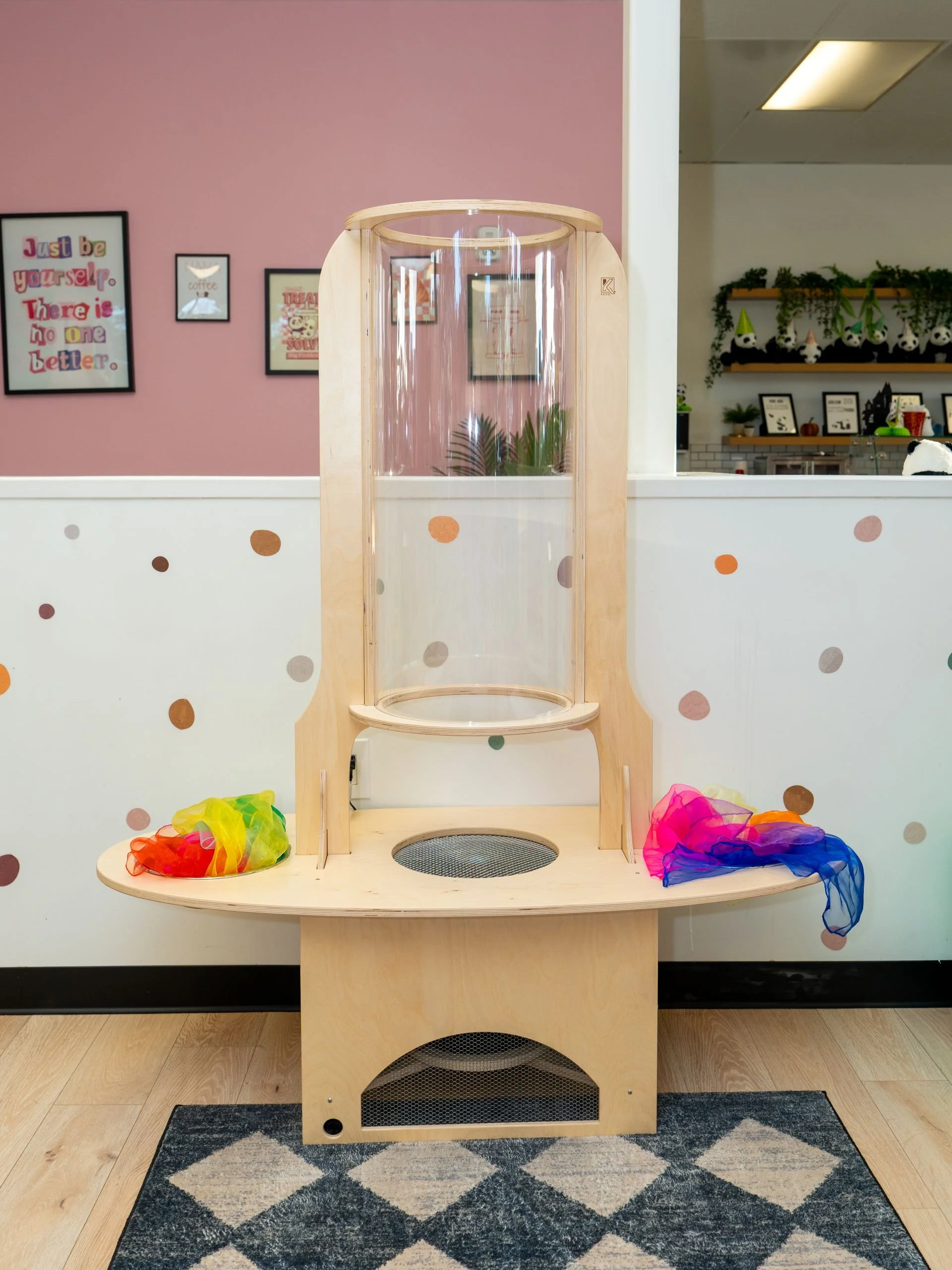 A wooden cat playhouse with a transparent tube and two colorful scarves on either side in a room with pink and white walls and framed wall art.