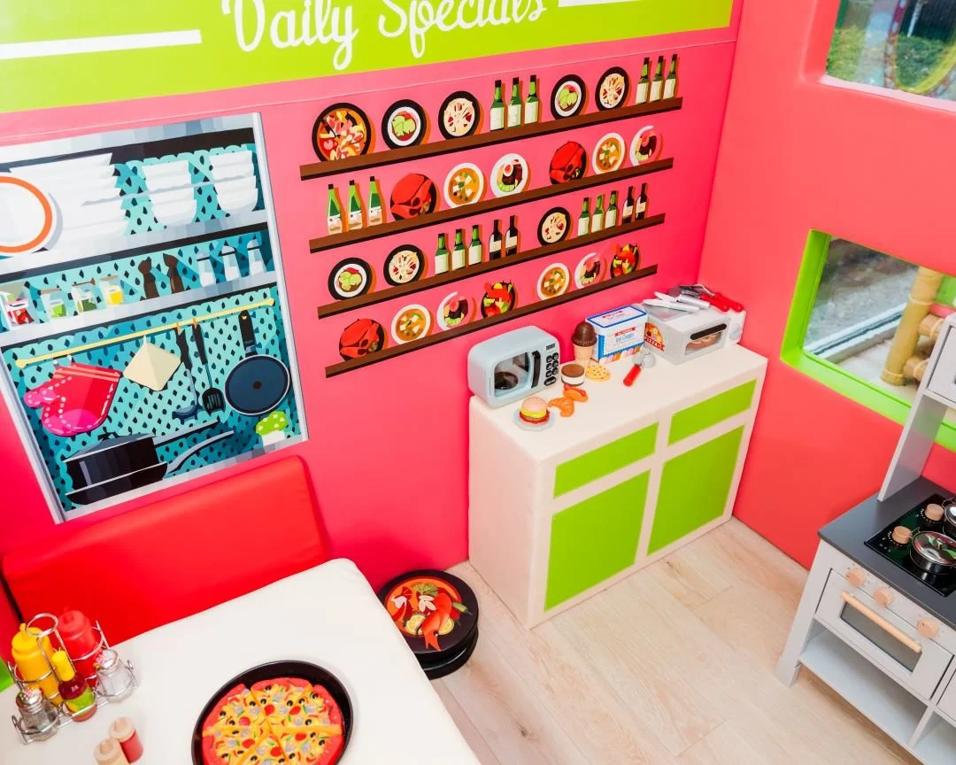 Toy kitchen with a white and green cabinet, a black stove, and a white counter with a microwave, toy food, and utensils. Pink walls decorated with colorful shelves filled with toy bottles and plates. A table with a tray of toy pizza slices, condiment