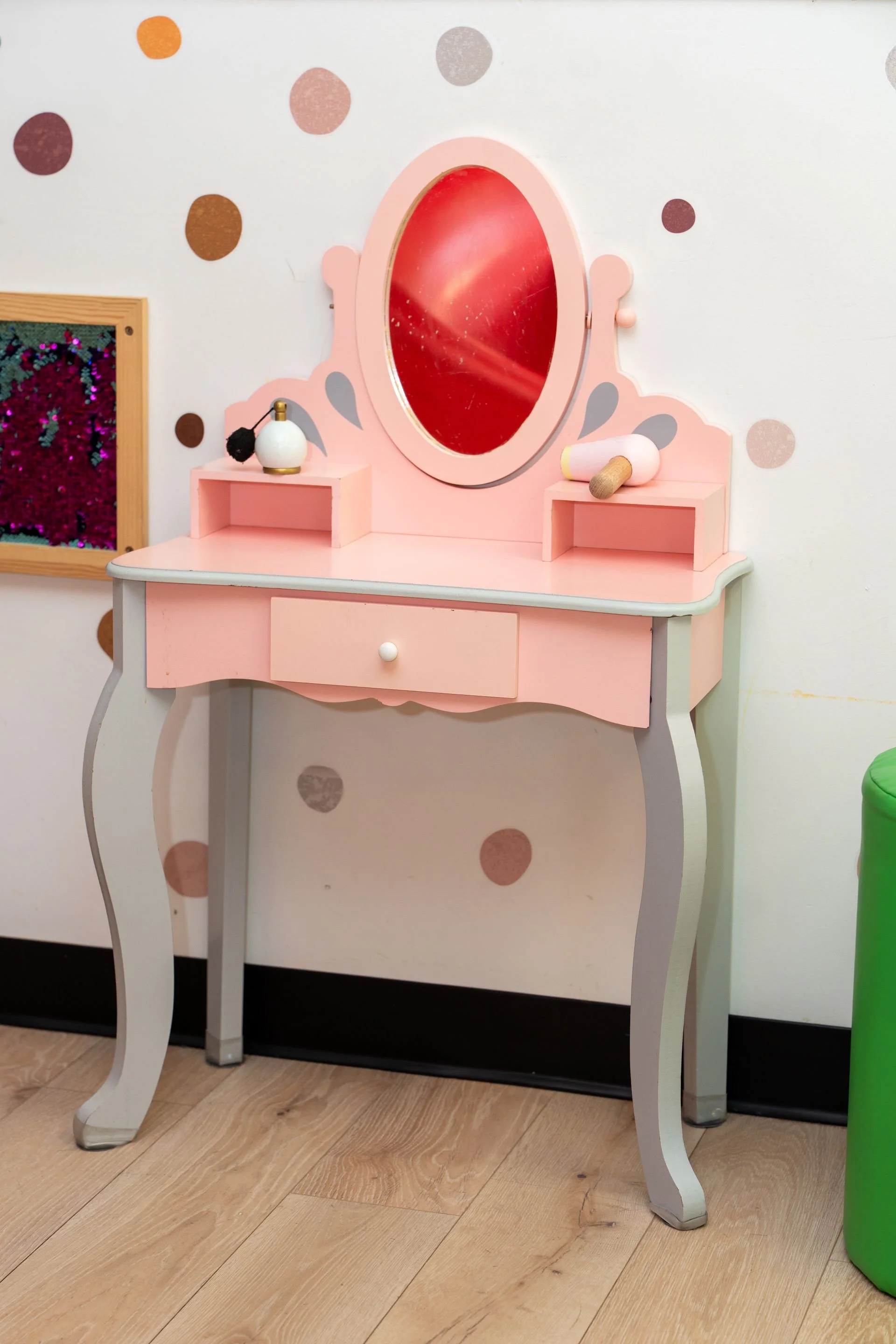 A pink and white vanity table with curved legs, a small drawer, and an oval mirror, situated against a wall with multicolored circular dots. The table has a small bottle and a rolled-up paper or scroll on top.