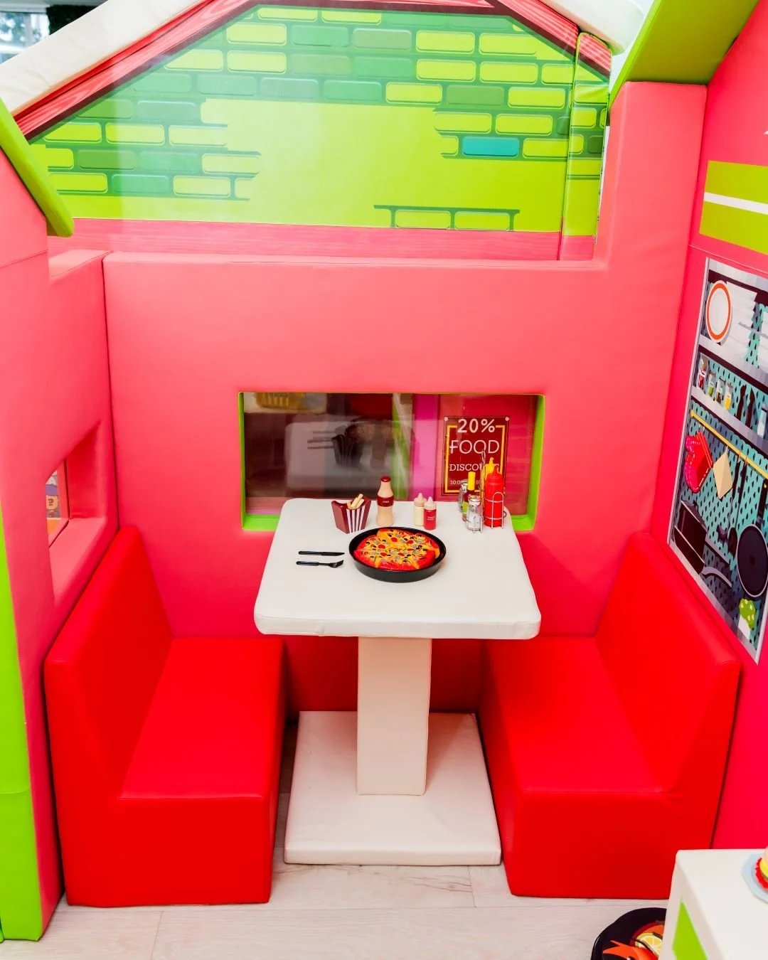 Colorful indoor play area with a small table set for pizza, surrounded by red and pink cushioned seats, featuring ketchup and mustard bottles, and a poster advertising 20% off on food.