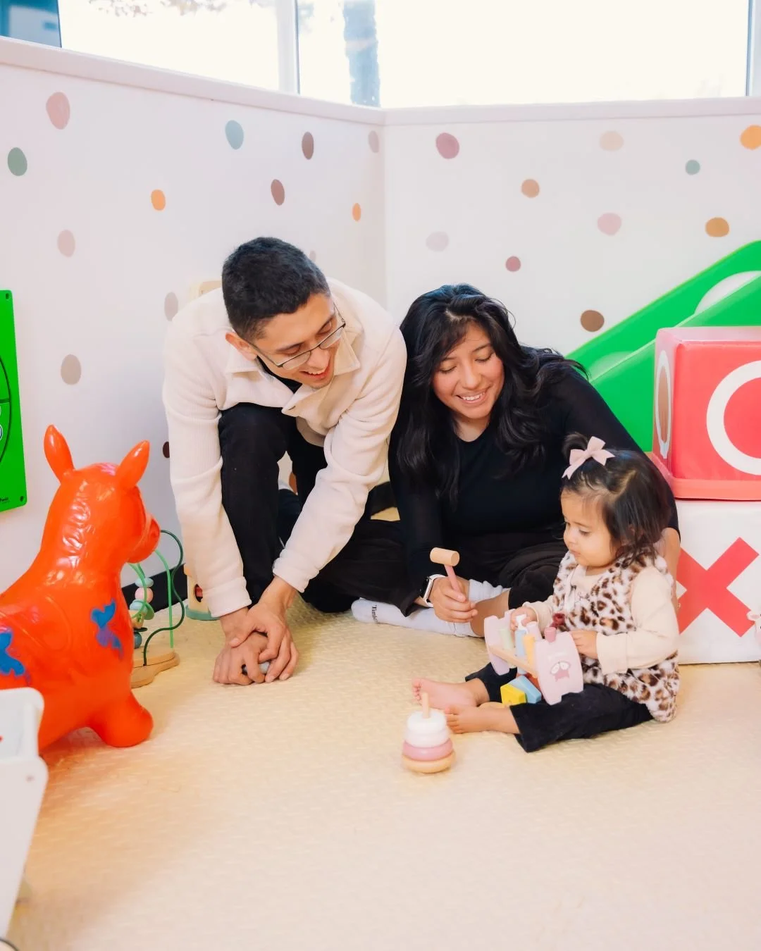 A family with a man, woman, and young girl playing together on a carpeted floor in a colorful indoor playground or playroom with toys and wall decorations.