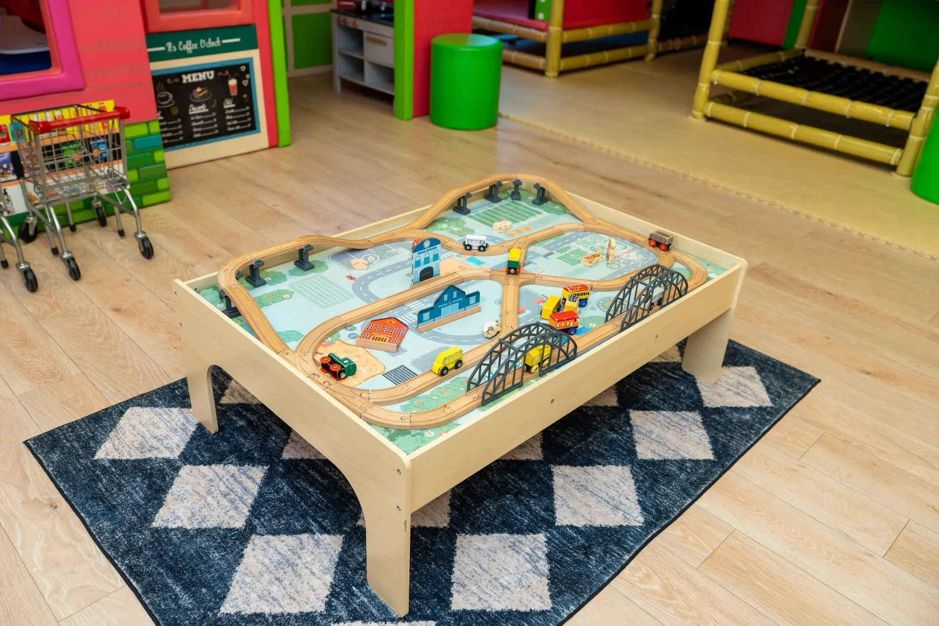 Children's train set with wooden tracks, train cars, and miniature buildings on a play table in a colorful indoor playroom.