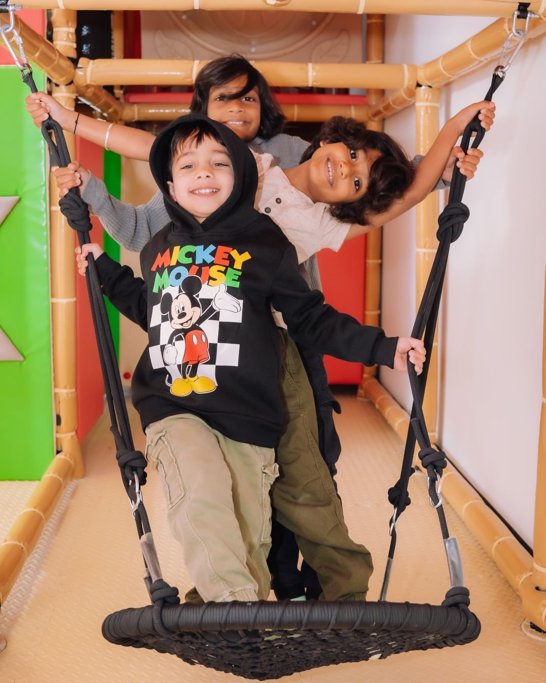 Three children playing on a indoor swing surrounded by colorful padded walls and bamboo-like structures.