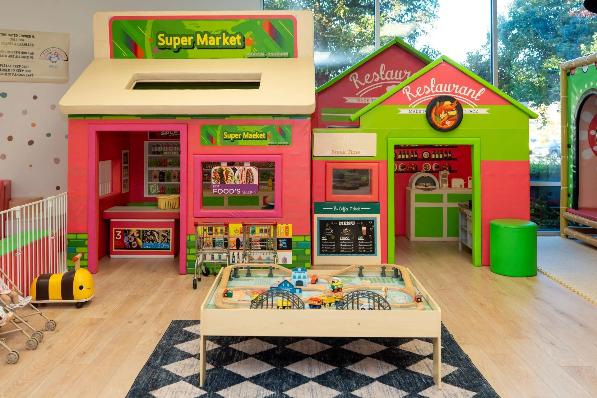 Children's play area with a pink and green pretend grocery store, a coffee shop, toy shopping carts, a train set in a wooden table, and toy strollers in the background.