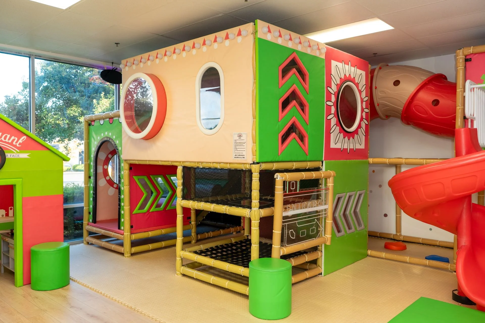 Indoor play structure with colorful tunnels, slides, and windows for children, in a room with large windows and a wood floor.