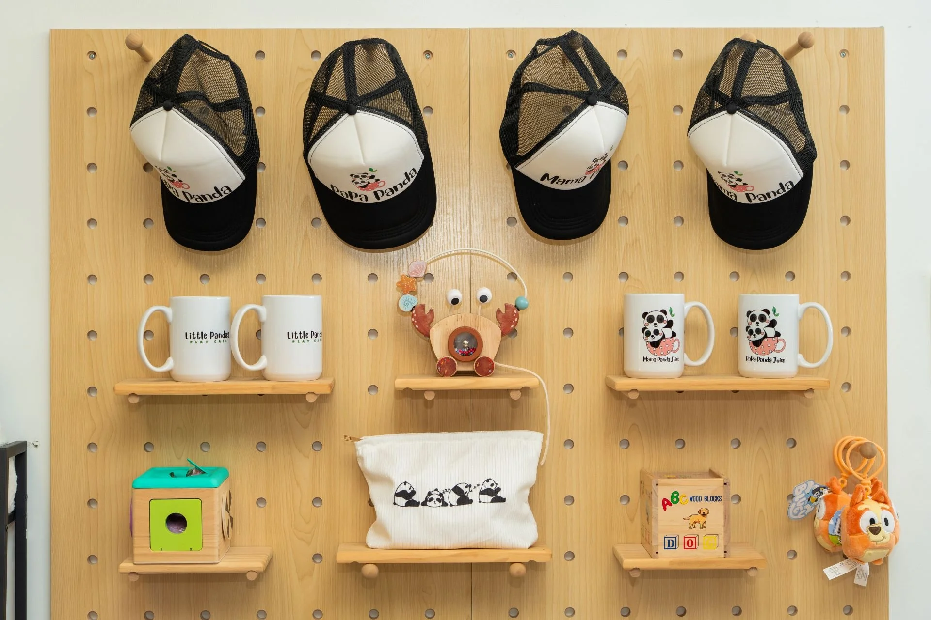 A wooden pegboard wall with four panda-themed caps hanging at the top. On the shelves below, there are two white mugs labeled 'Little Panda Play Cafe,' two mugs with panda graphics labeled 'Mama Panda Juice' and 'Papa Panda Juice,' a wooden panda fig