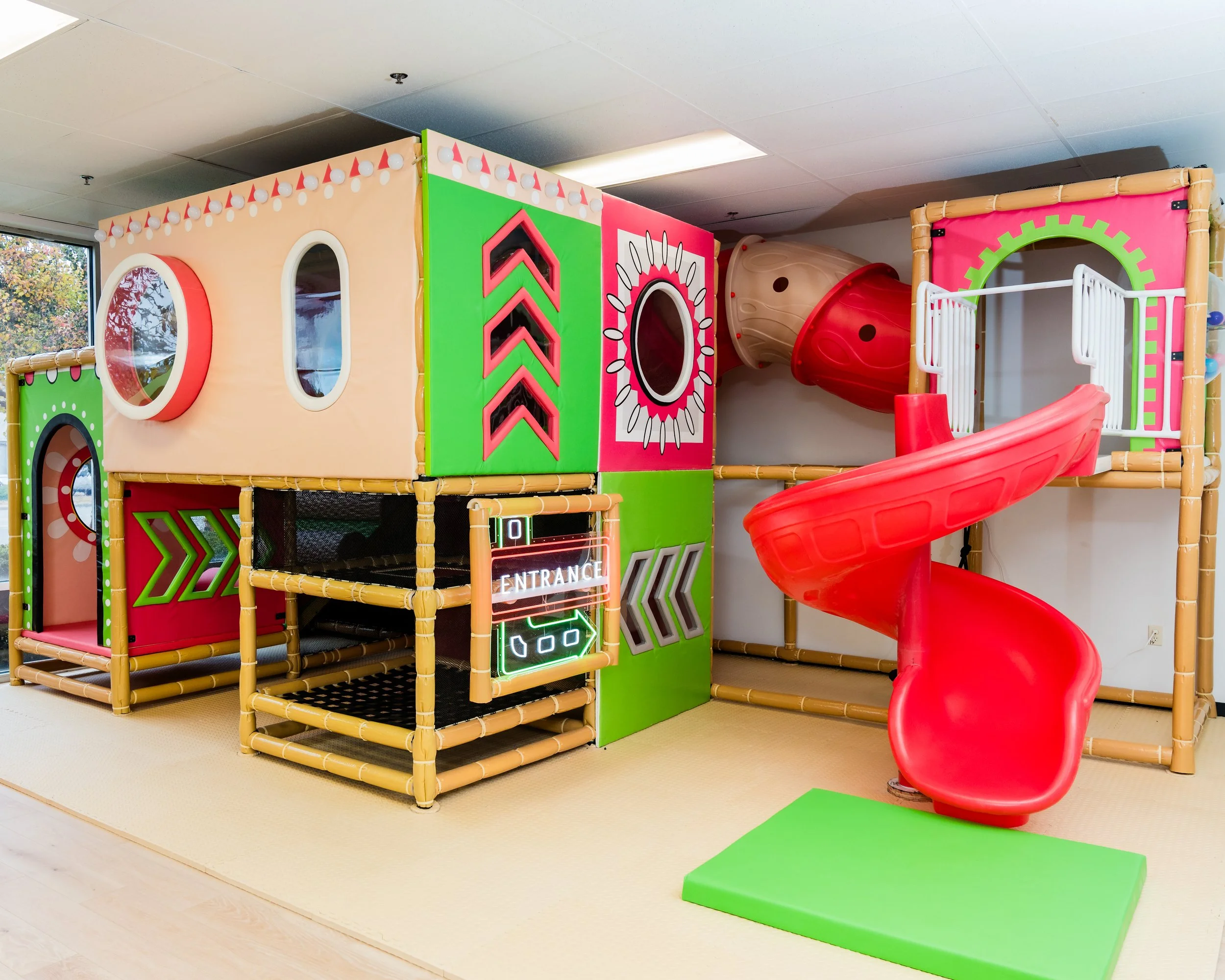 Indoor play structure for children featuring colorful sections, a red spiral slide, tunnel, and climbing areas, with a neon 'Entrance' sign.