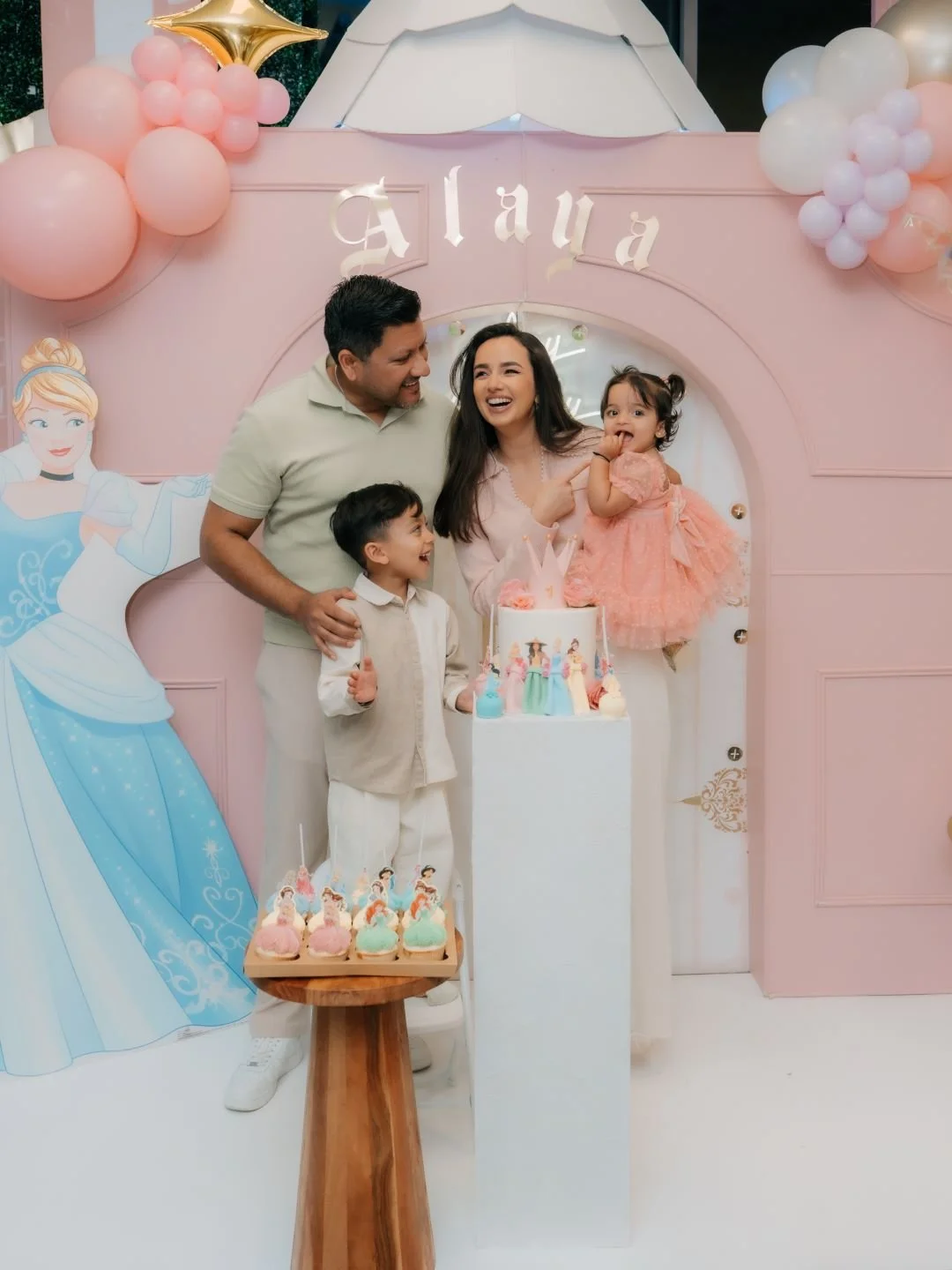 Family celebrating a birthday party with princess-themed decorations, cake, and cupcakes, in front of a pink backdrop with balloons and princess illustrations.