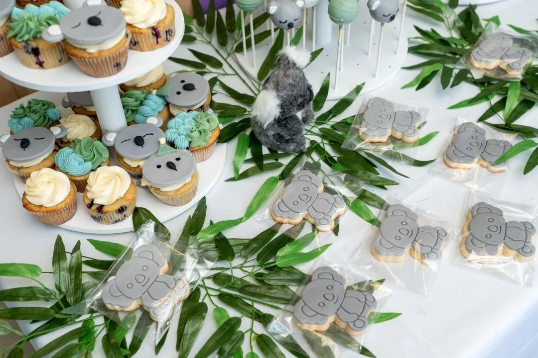 Decorated cupcakes with koala faces and eucalyptus leaves, koala plush toy, and koala-shaped cookies wrapped in plastic on a white table for a themed party.