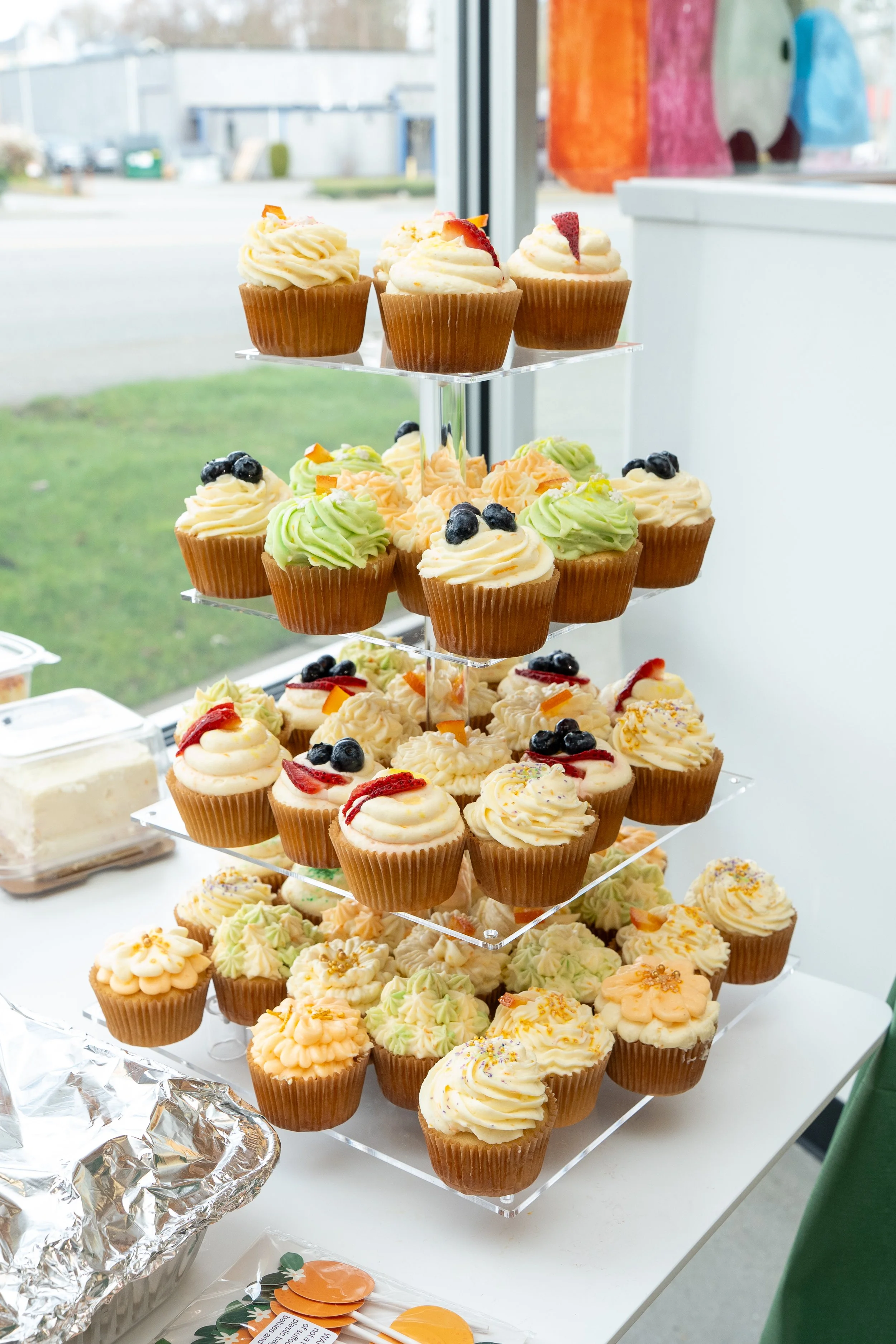 Tiered display of assorted cupcakes with various frosting and decorative toppings, set up near a window.