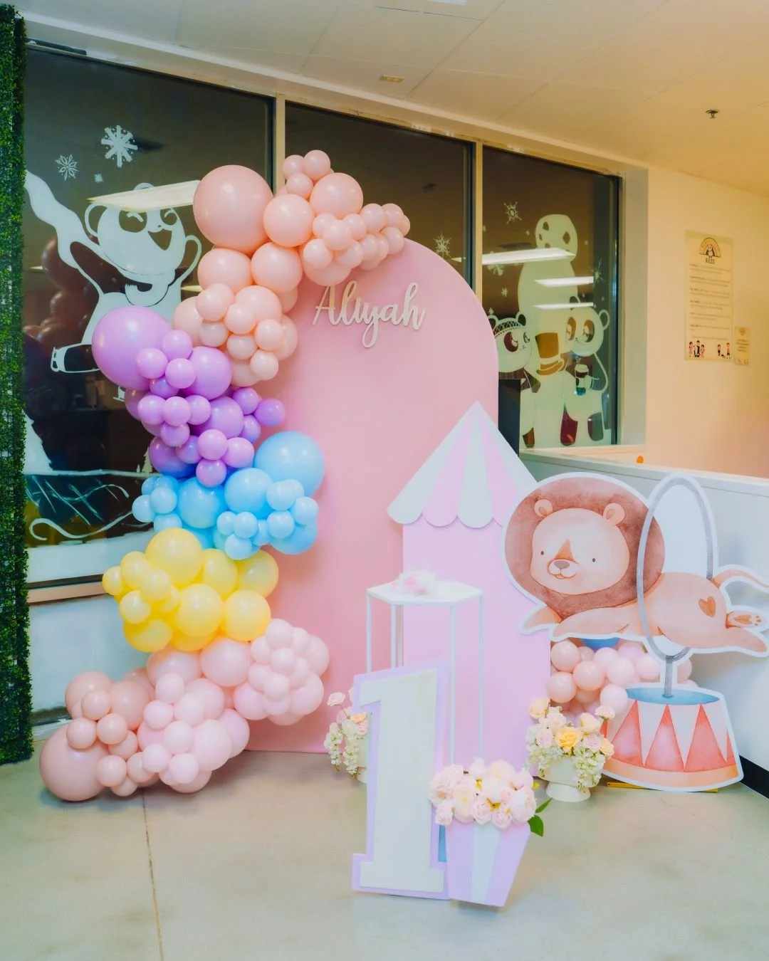 Decorations for a first birthday celebration for a girl named Alyah, featuring pink, purple, blue, yellow balloons, a pink backdrop with her name, a large number one with flowers, and cartoon animal cutouts including a lion with a hat and musical not