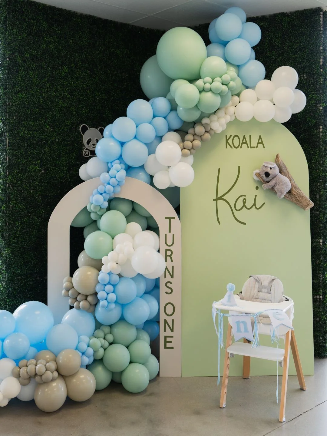 Decorative display for a baby shower with a pastel balloon arrangement, plush koala toy attached to a wooden branch, and a high chair with a banner that says 'one'.