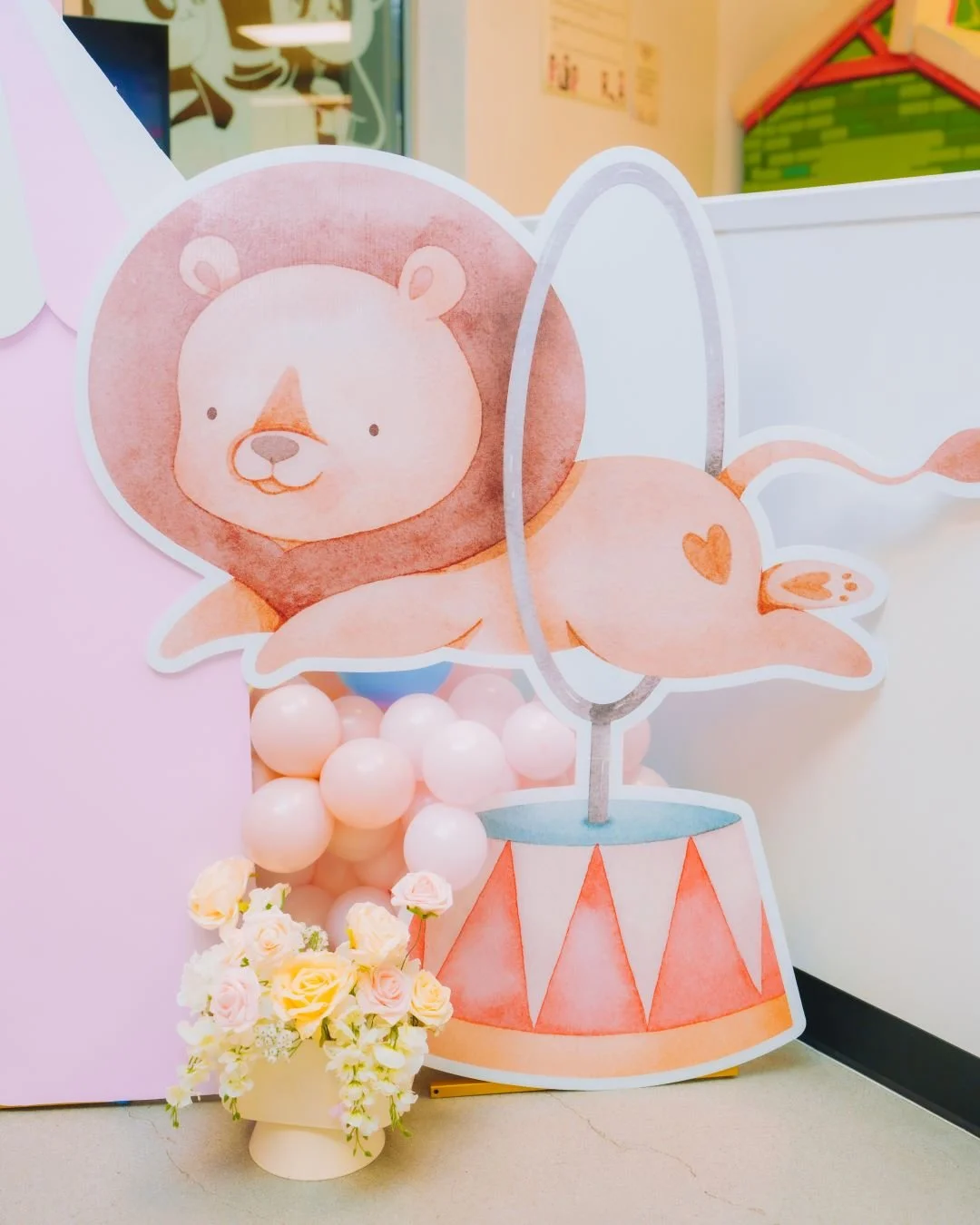 A decorative cutout features a cute illustrated lion with a round face and a lion's mane, lying on a circus-style platform. The display is complemented by pink and white balloons and a small bouquet of pastel-colored roses in front.