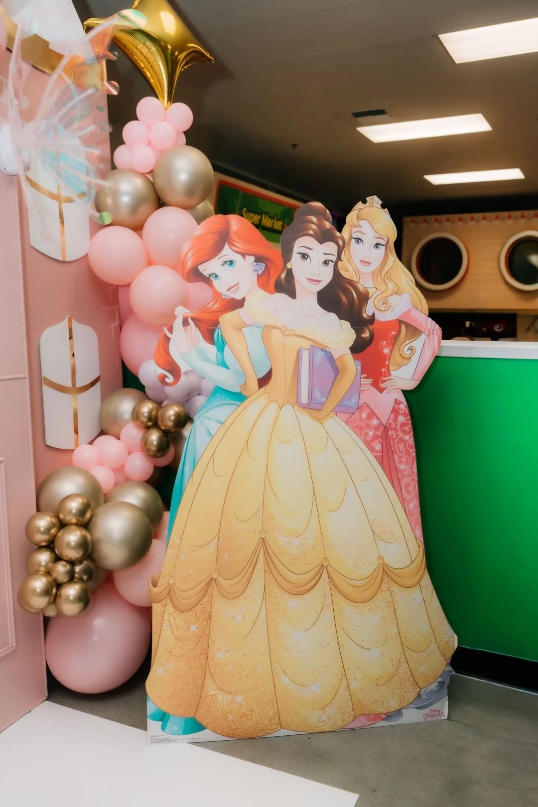 Decorative display with cutouts of Disney princesses Ariel, Belle, and Aurora, surrounded by pink, gold, and white balloons, at a festive event.
