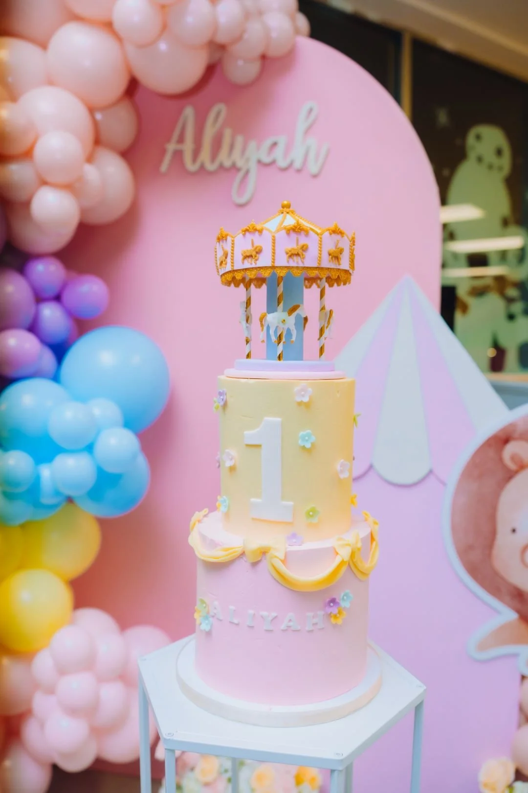 A pastel-colored, three-tiered cake decorated with flowers, a number one, and a carousel topper, celebrating a first birthday for Alyah, with a pink background and balloon decorations.