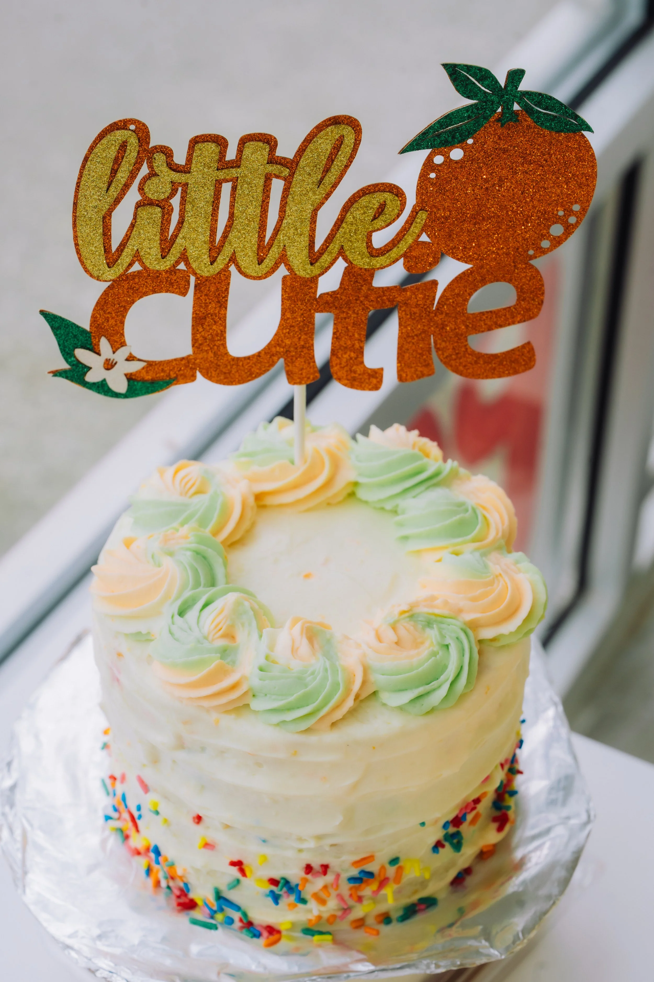 A birthday cake decorated with pastel-colored swirls of frosting around the top edge and rainbow sprinkles on the side, topped with an orange and green "little queen" cake topper shaped like a citrus fruit with leaves and a white flower.