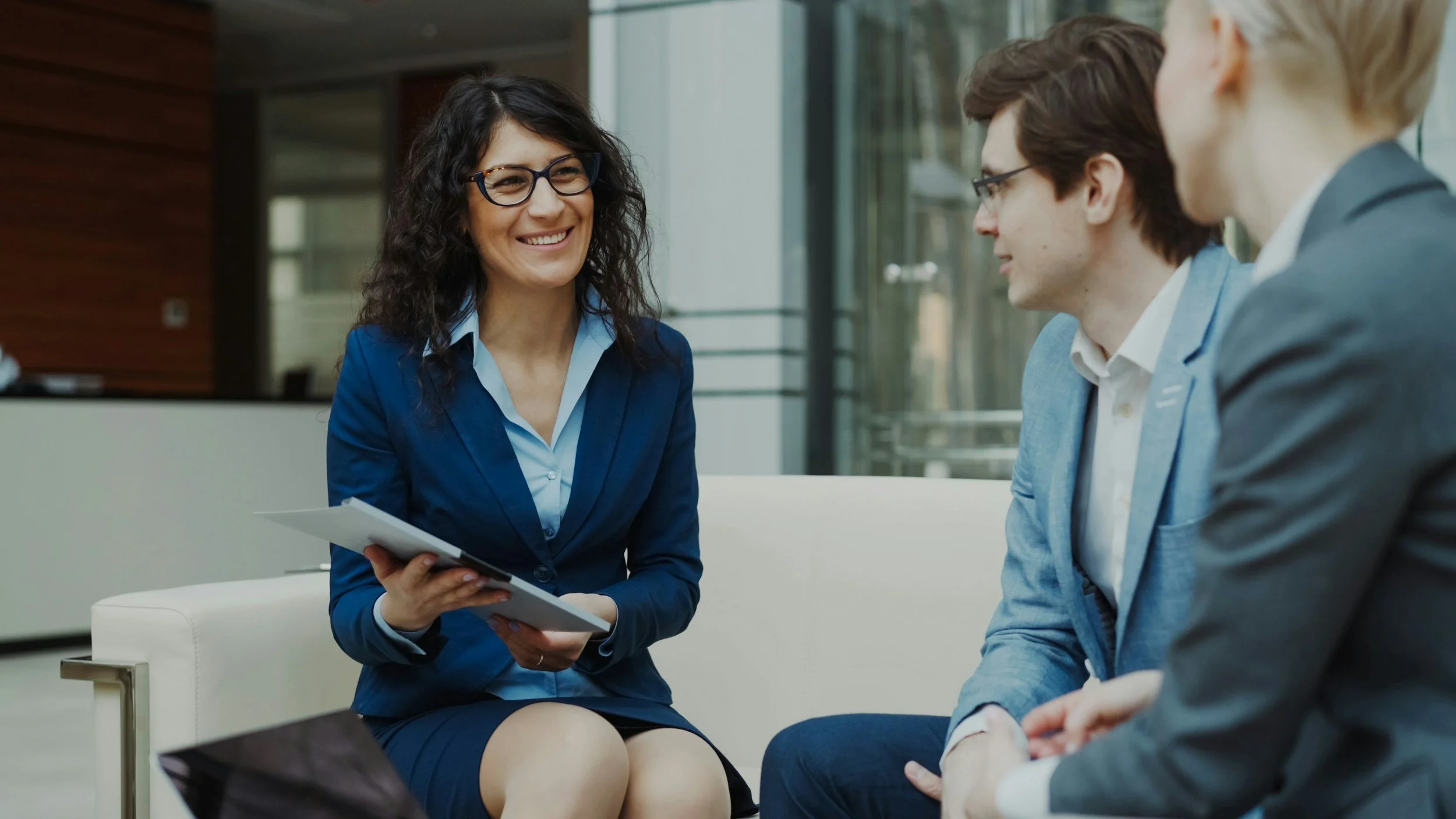 A woman with dark curly hair and glasses smiling while holding papers, engaged in conversation with two men in suits in a modern office setting.