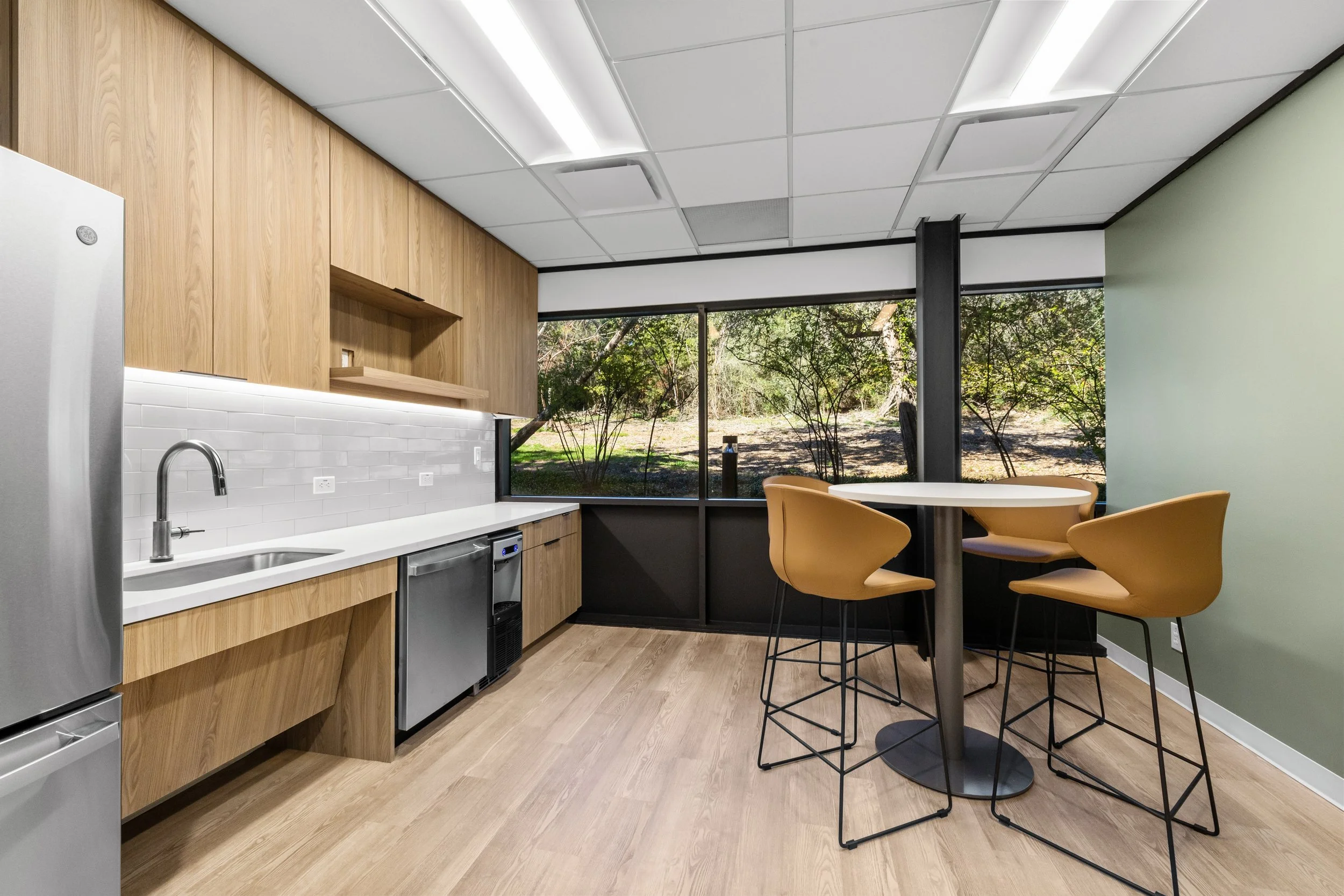 Office breakroom construction by Romann Construction in DFW, showcasing light wood cabinetry, stainless steel finishes, and large windows.